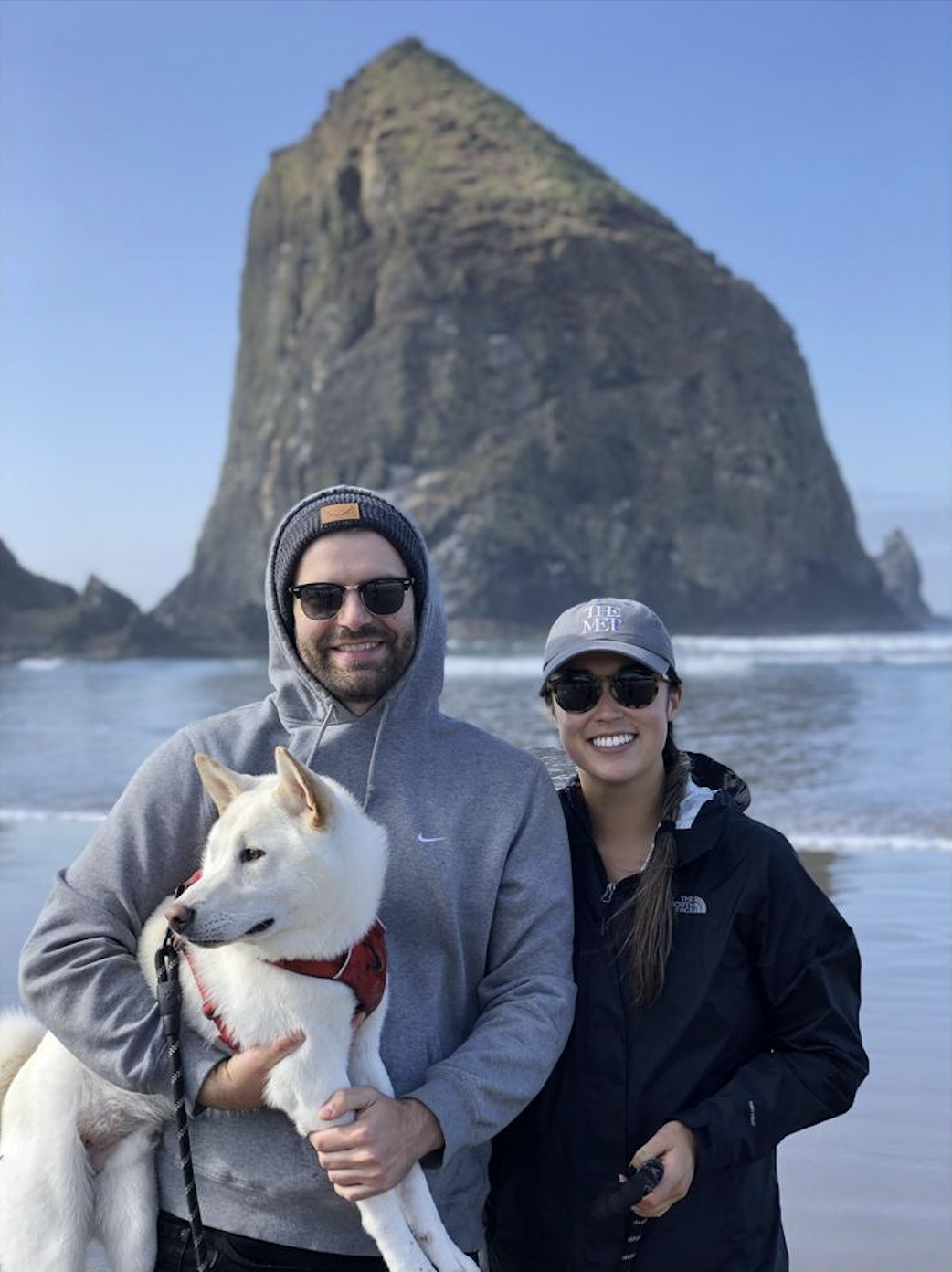 A smiling man and woman in outdoor clothing stand on a beach with a large rock formation in the background. The man is holding a white dog with a red harness.