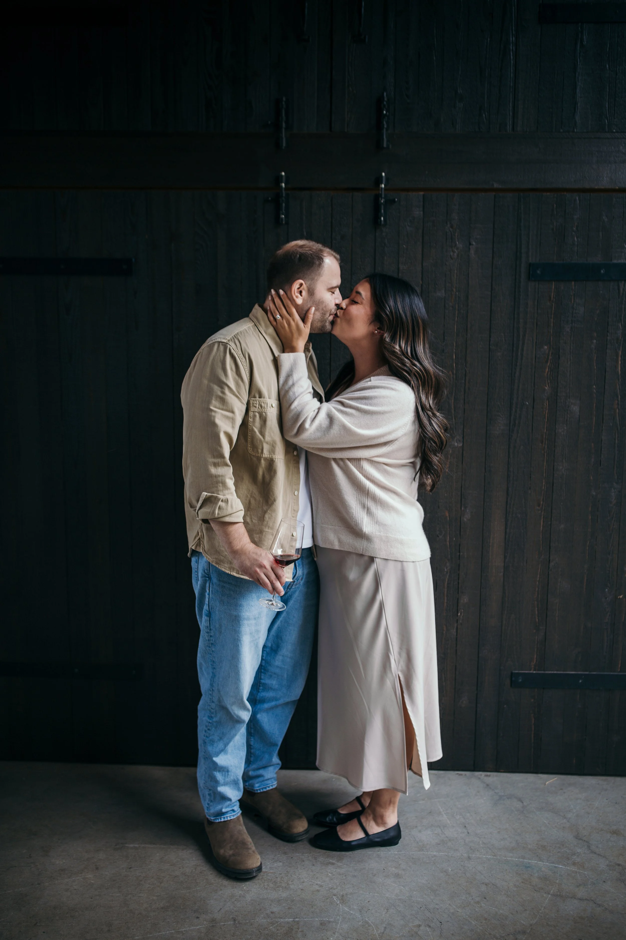 A couple kissing, woman holding wine glass, standing against dark wooden wall.