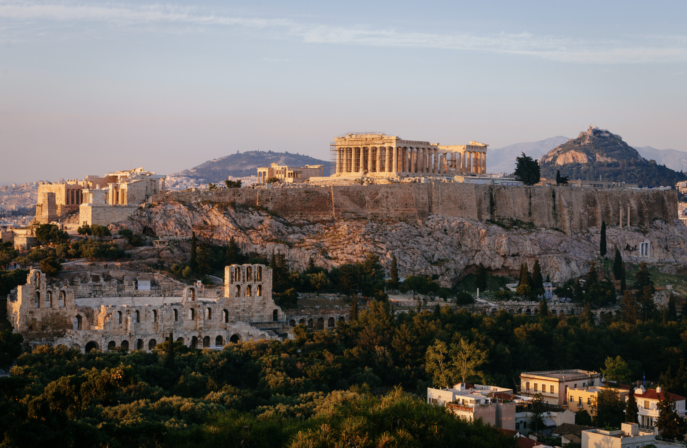Sunset view of the Acropolis in Athens, Greece, featuring the Parthenon atop the rocky hill, with ancient ruins and lush greenery in the foreground.
