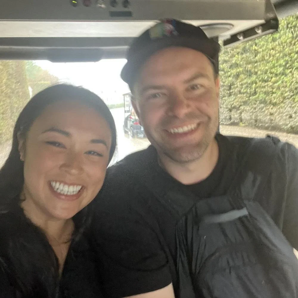 A woman and a man smiling and taking a selfie together inside a vehicle, with greenery and a road visible through the windows in the background.