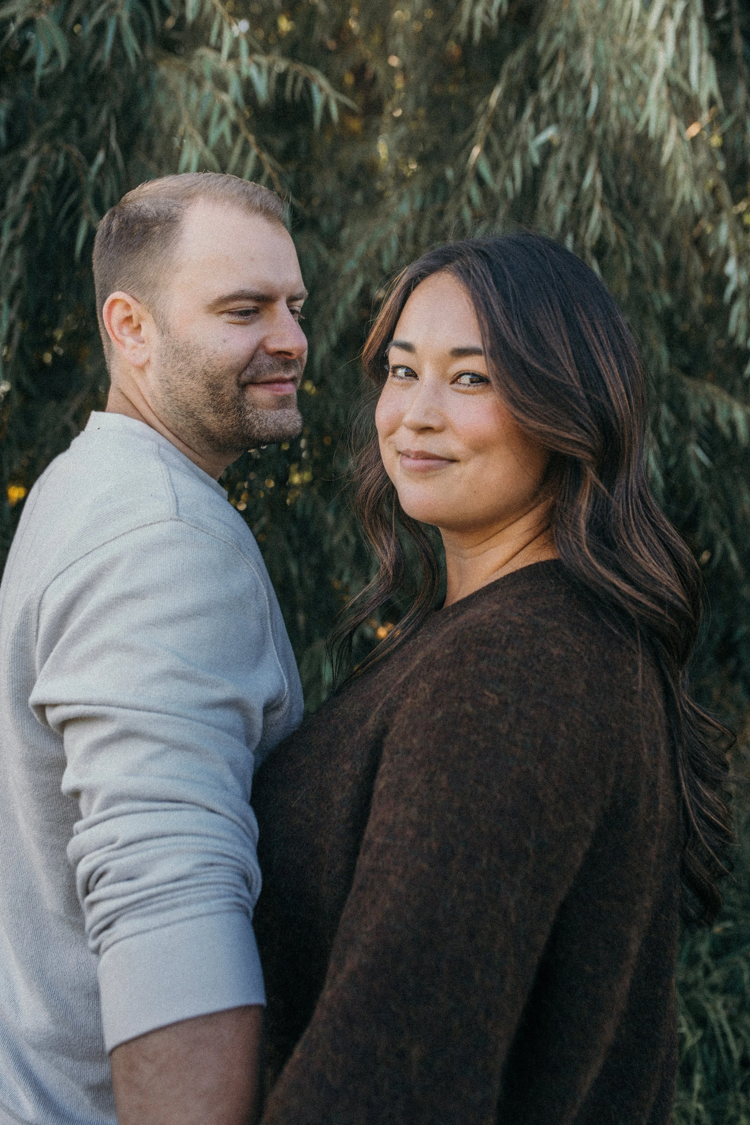 A man and woman standing outdoors near trees, looking at the camera with the man smiling slightly and the woman smiling gently.