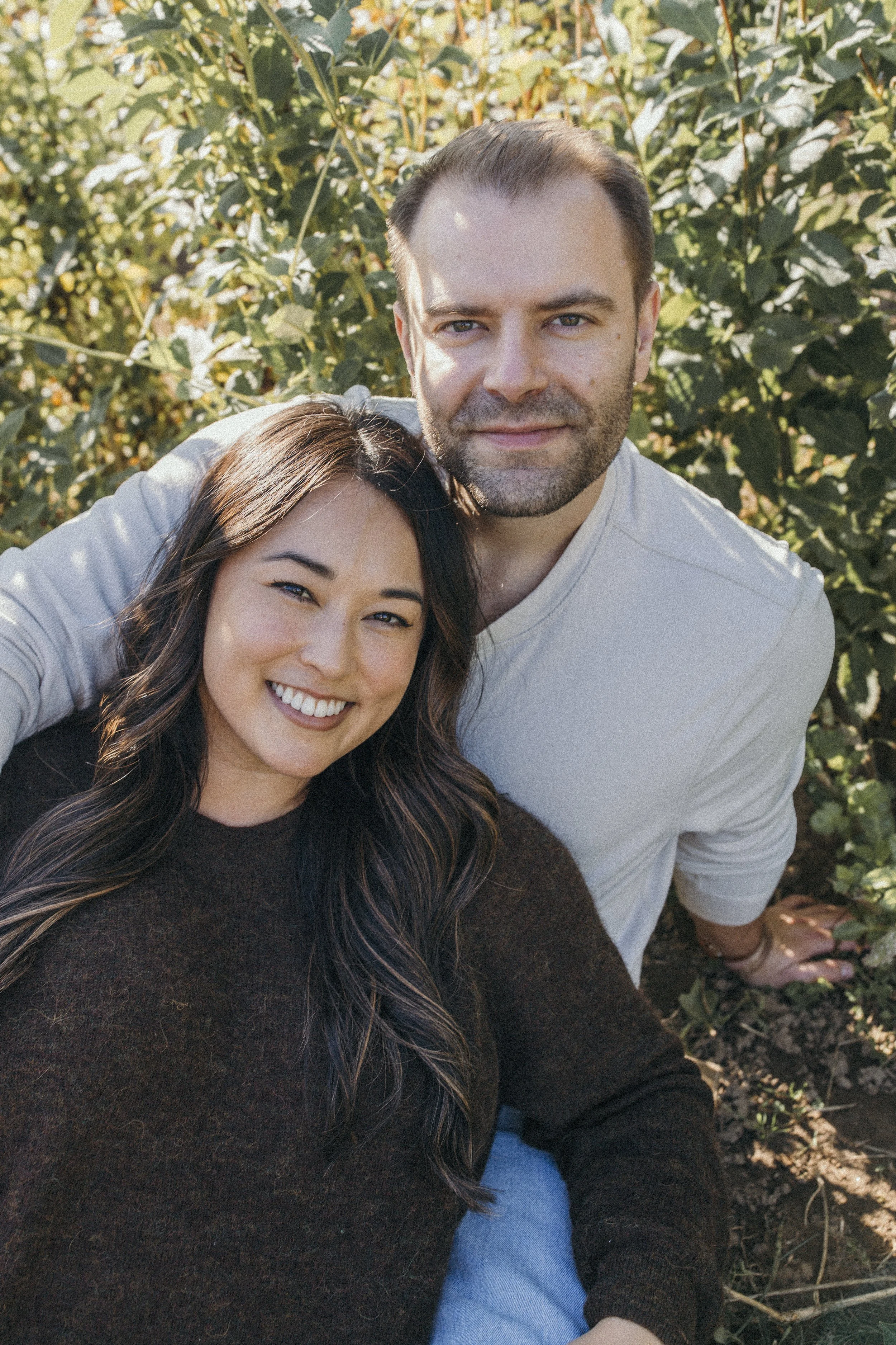 A smiling woman with long wavy dark hair and a man with short brown hair and a beard sitting outdoors among green bushes. The woman is wearing a dark sweater, and the man is in a light-colored shirt. They are close together, smiling and looking at th