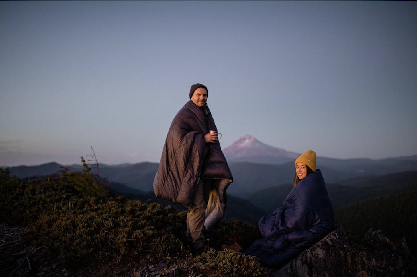A man and woman wrapped in sleeping bags on a mountain top during sunset, mountain landscape in the background, woman wearing a yellow beanie.