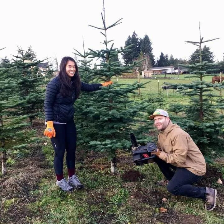 Two people, a woman and a man, are in a Christmas tree farm. The woman is standing and smiling, touching a small fir tree. She is wearing a navy jacket, black pants, and gray shoes. The man is squatting next to her, holding a power tool, and smiling.