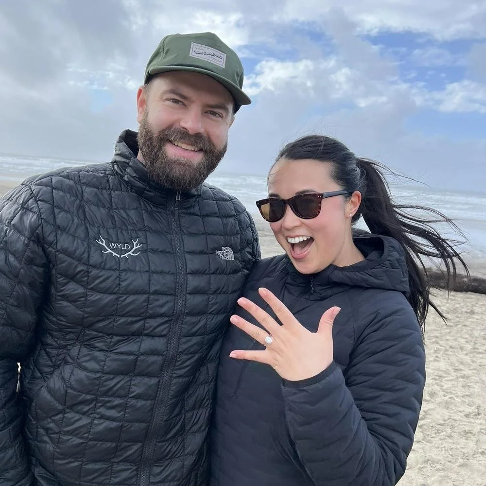 A smiling couple on the beach, with the woman showing off an engagement ring. The man is wearing a black quilted jacket and a green cap, while the woman is wearing sunglasses and a black jacket. The ocean and cloudy sky are visible in the background.