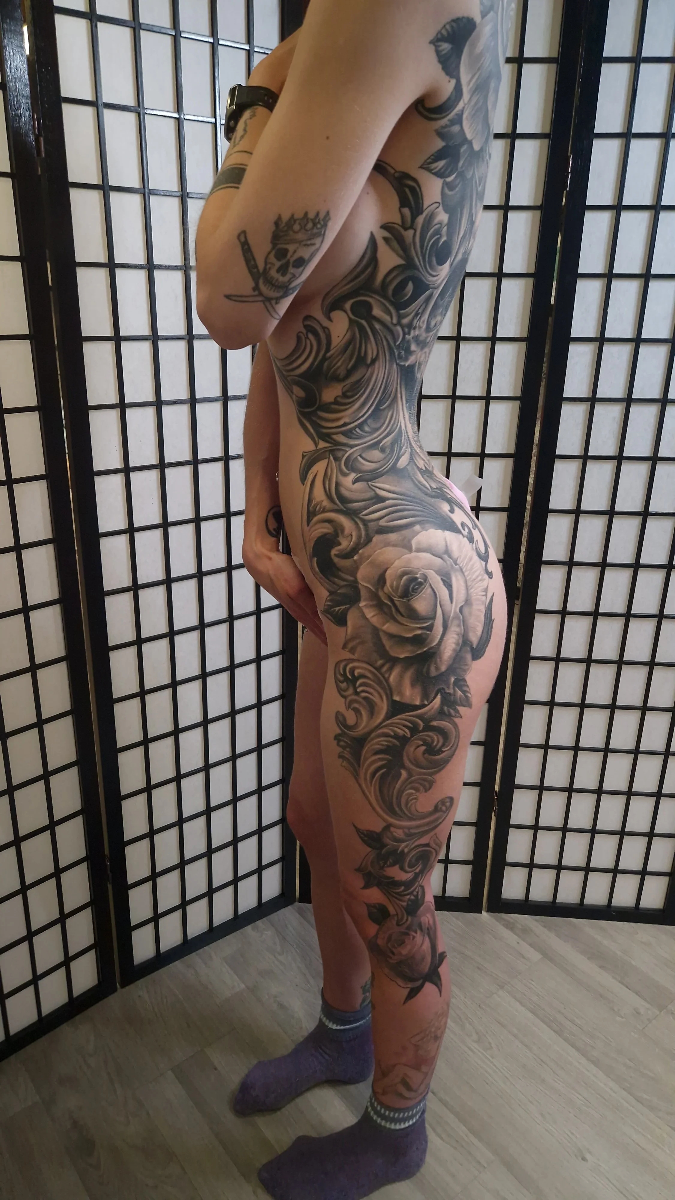 A person with extensive black and gray tattoos, including a skull with a crown and sword on the arm, a large floral design on the torso, and roses on the leg, standing in front of a black grid-style room divider.
