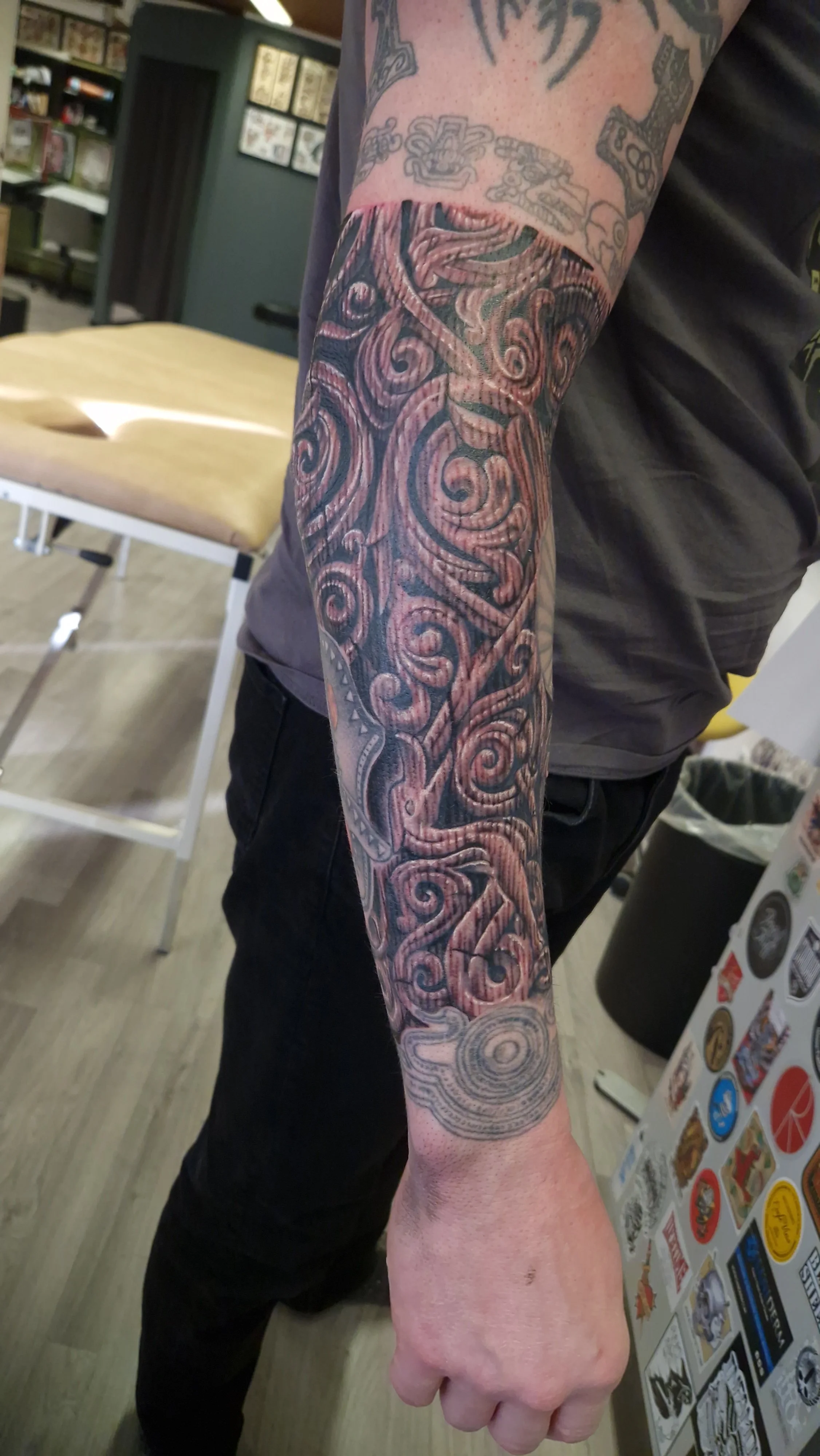 A person's arm with a detailed black and grey Polynesian-style tattoo sleeve featuring intricate swirling and geometric patterns.