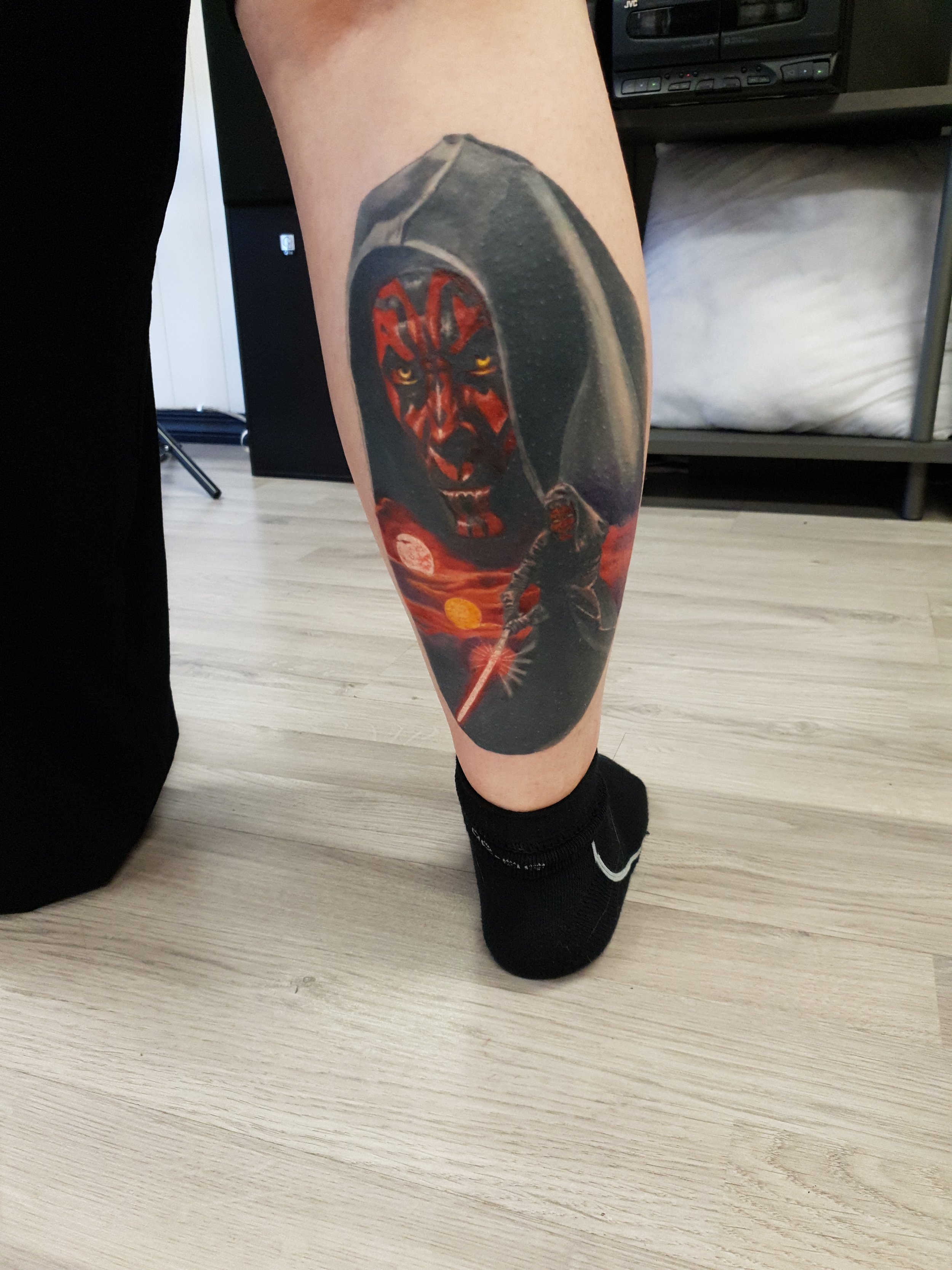 Tattoo of Darth Maul from Star Wars on a person's lower leg, with a background of a sci-fi scene including a figure wielding a red lightsaber near lava and a double sunset.