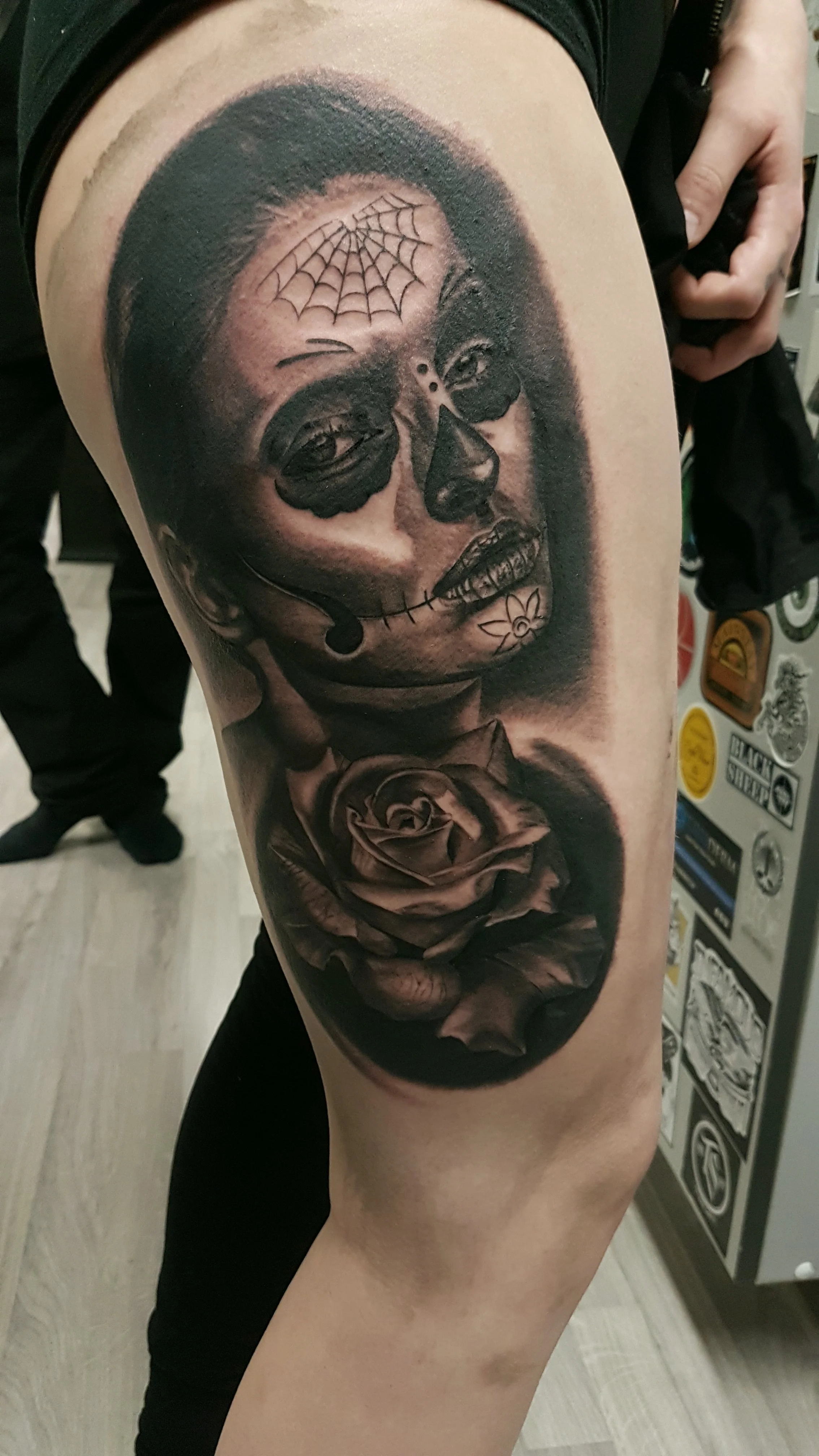 A black and gray realistic tattoo of a woman's face with day of the dead makeup, a spider web on forehead, a flower on cheek, and a rose below. The woman has earrings and appears to be winking.