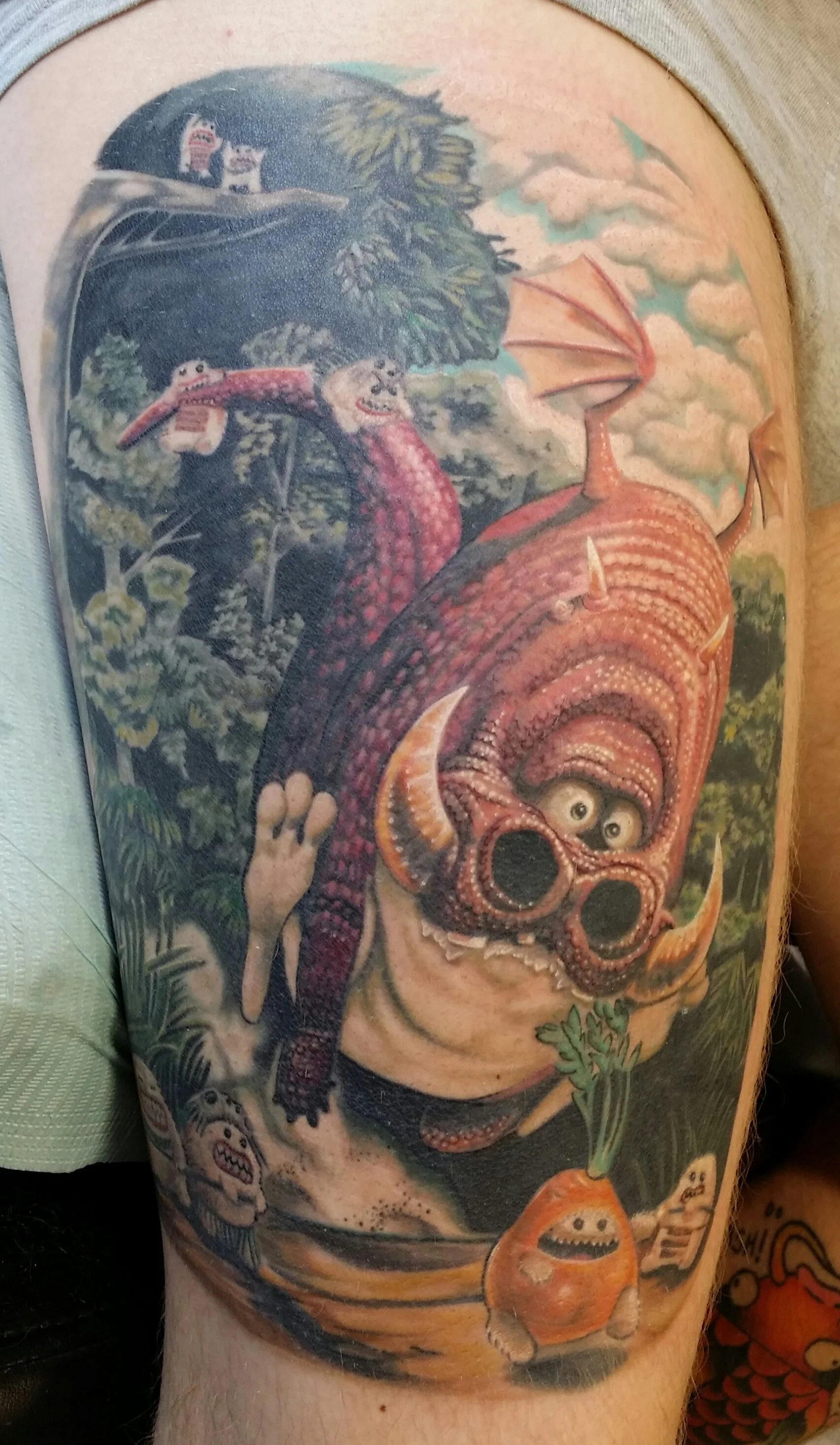 Colorful tattoo featuring a large, cartoonish monster with a horned head, big eyes, and a wide mouth, carrying smaller monsters. The background shows trees, clouds, and a flying creature with wings.