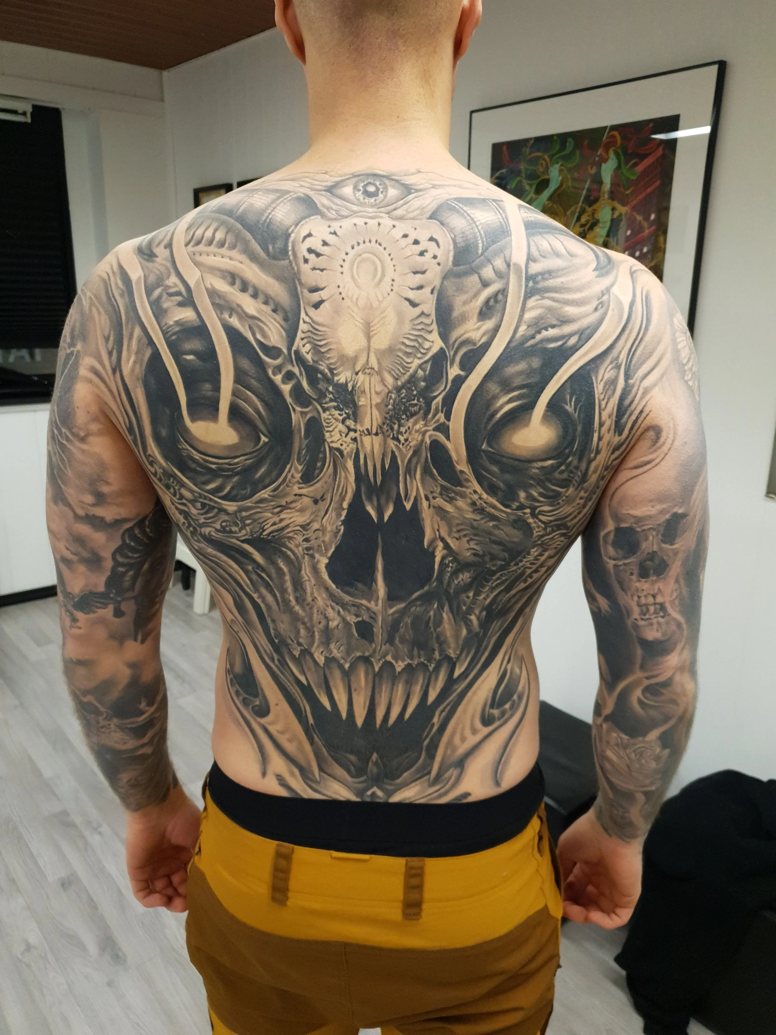 A man with a large black and gray tattoo covering his back, featuring a skull with sharp teeth, intricate patterns, and a detailed face with an eye at the top of his back, in a room with framed artwork on the wall.