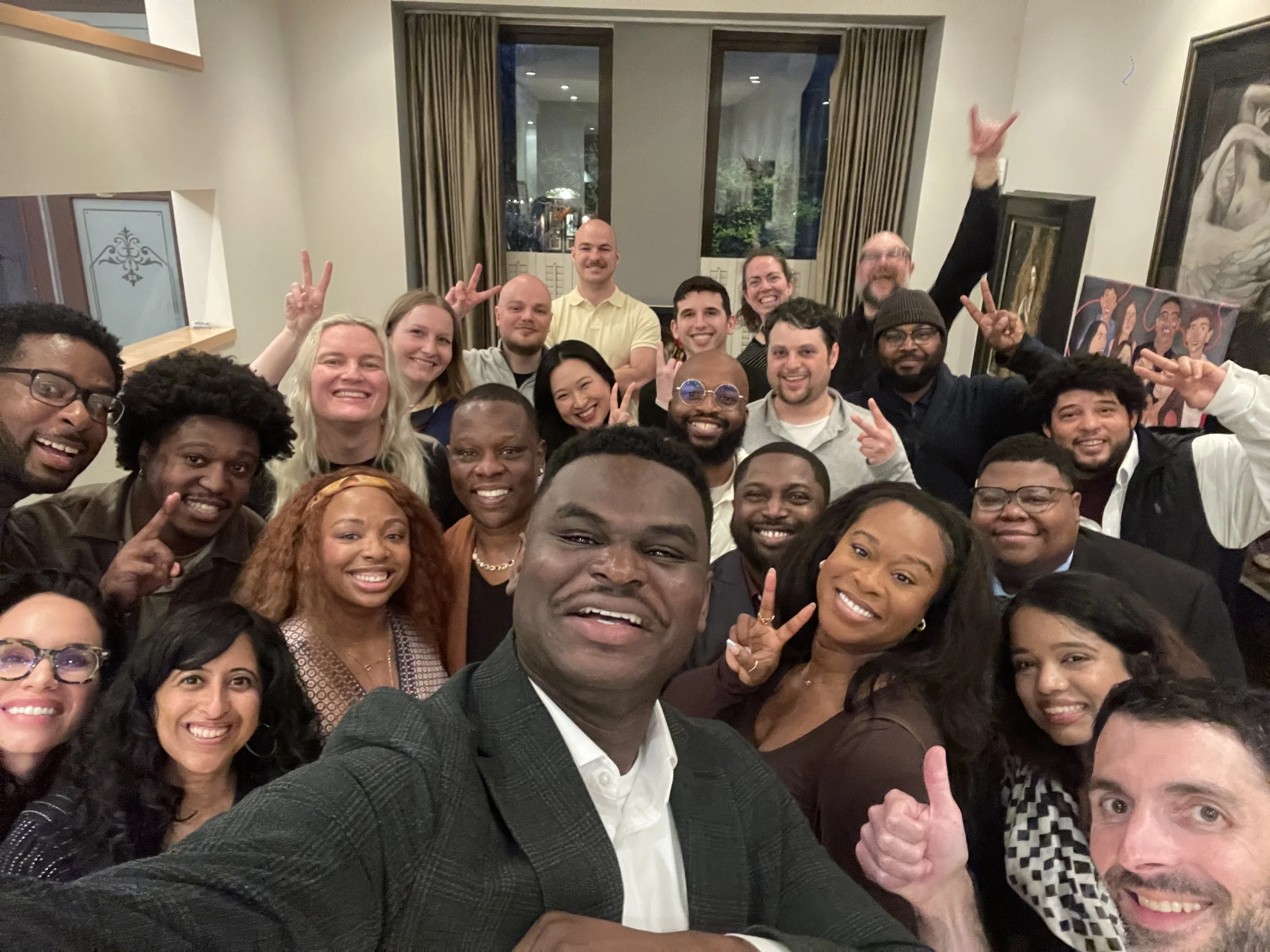 A group of about 30 diverse smiling people gathered in a room for a selfie, with some making peace signs and thumbs-up gestures. Richard (the candidate) is front and center smiling as he takes the photo.