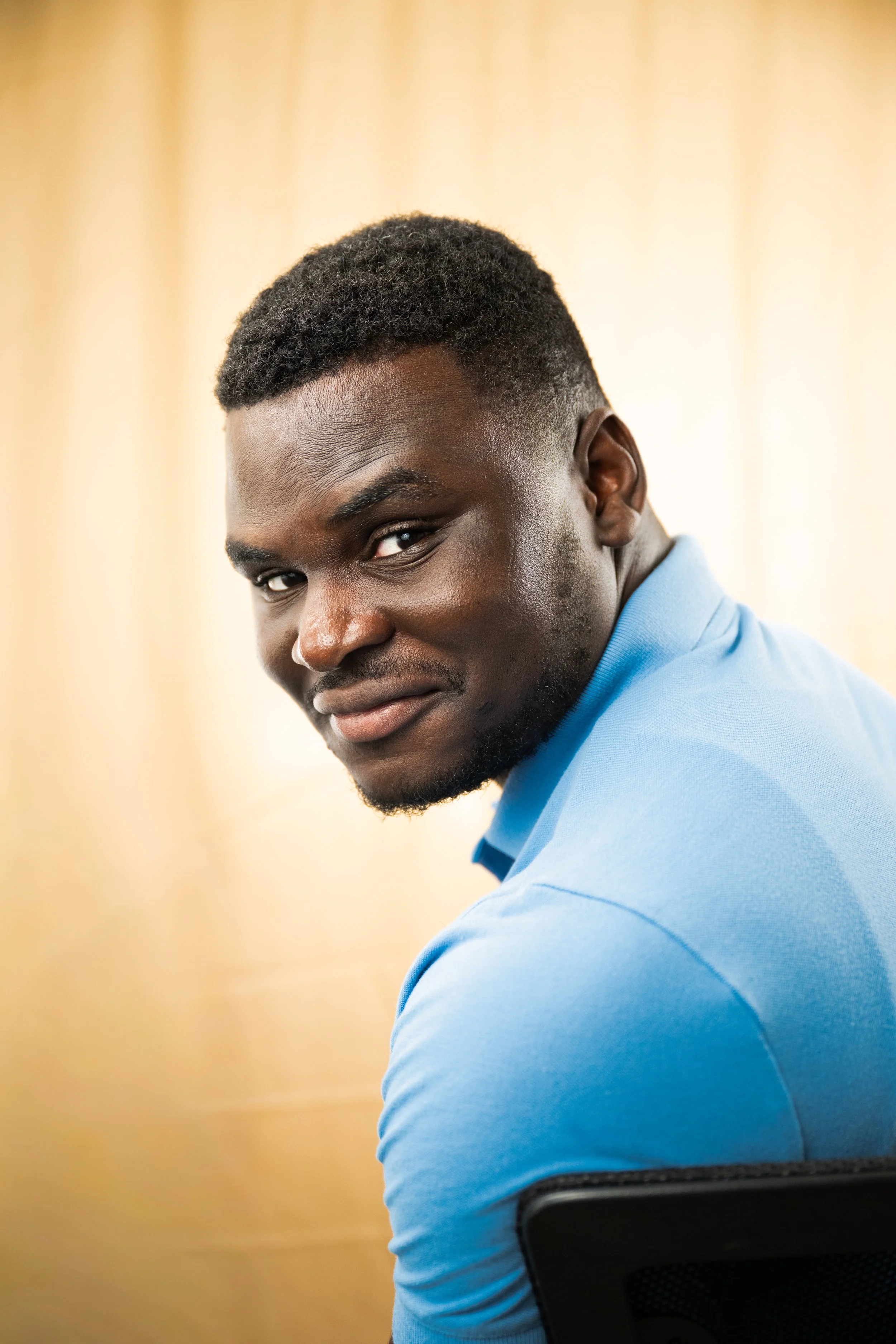 A young man with dark skin and short curly hair wearing a blue polo shirt, looking over his shoulder and smiling while sitting in an indoor setting with a blurred beige background.