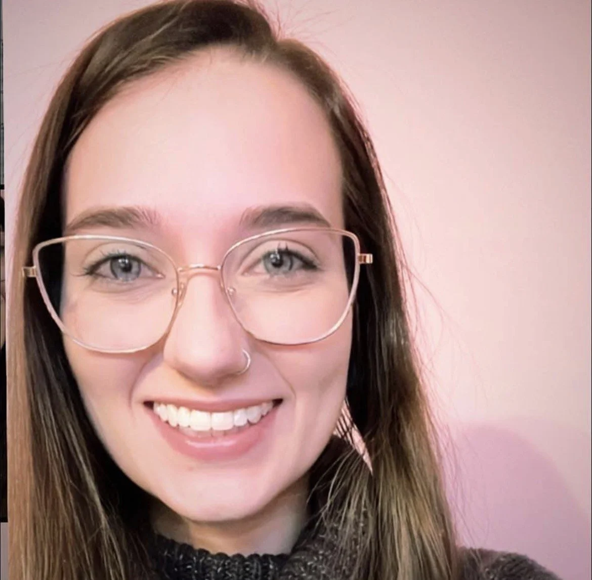 Headshot of Stephanie smiling, with long brown hair, rose-gold wire-frame glasses, and a dark knit sweater against a soft pink background.