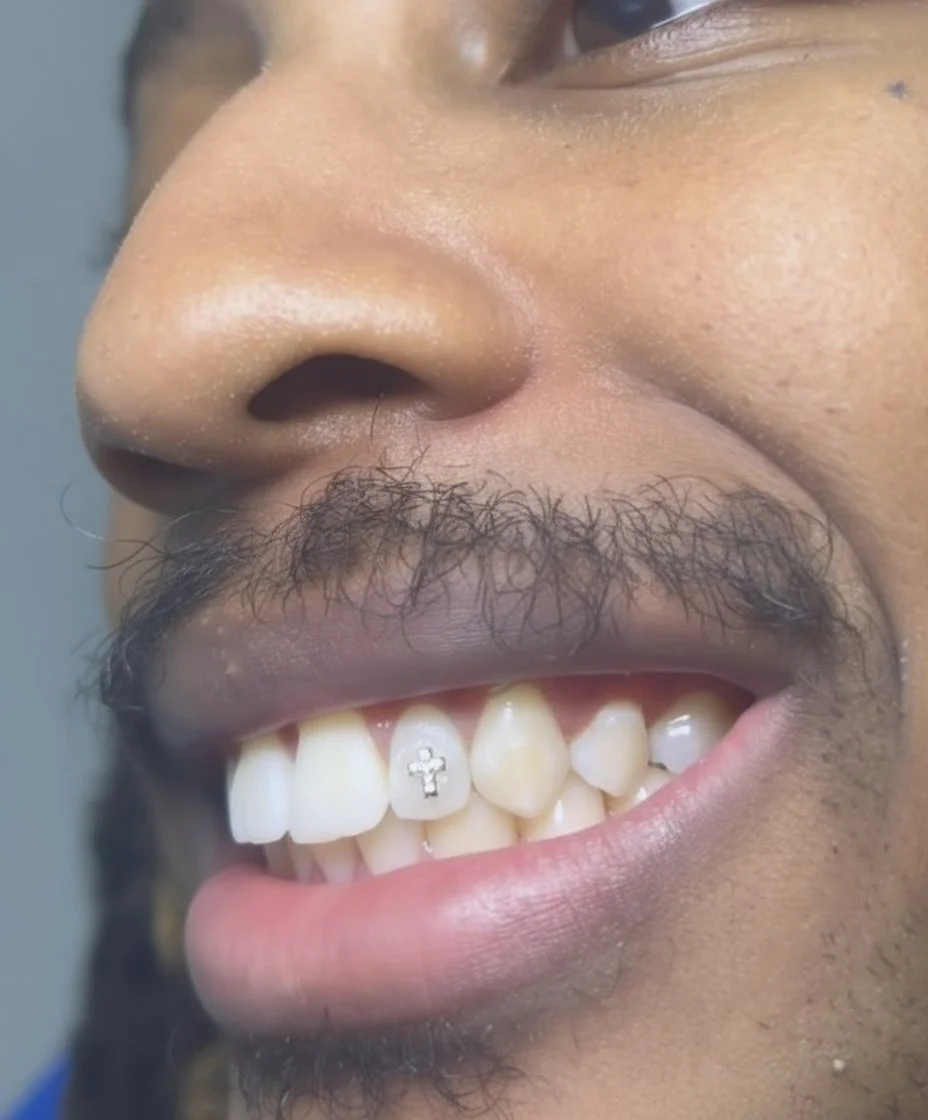 Close-up of a smiling person's face, showing nose, mustache, and teeth. The person has a piercing with a cross charm on one tooth.