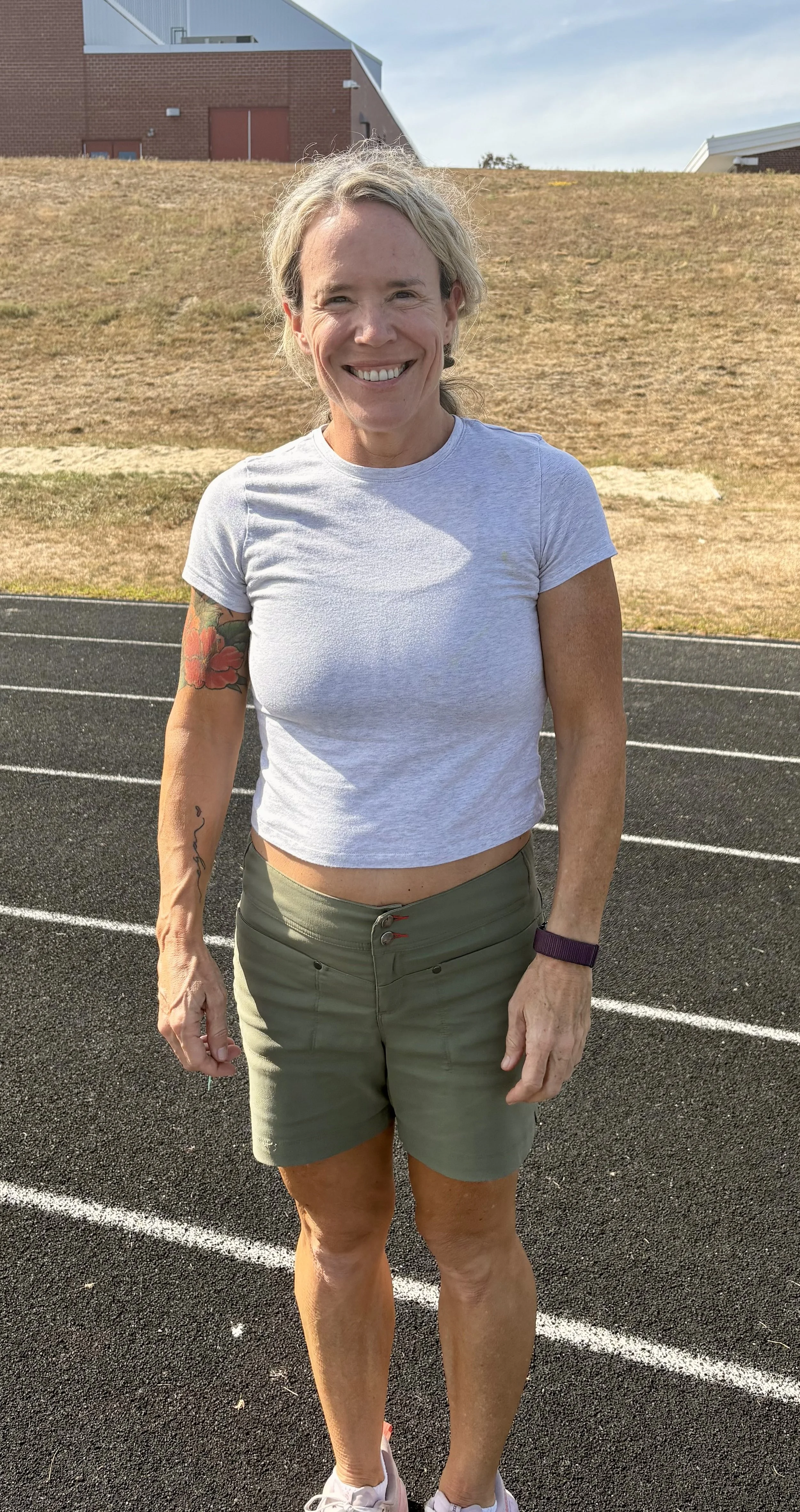 Woman standing on a running track, smiling, wearing a white t-shirt and green shorts, with tattoos on her left arm, in an outdoor setting with a building and grass in the background.
