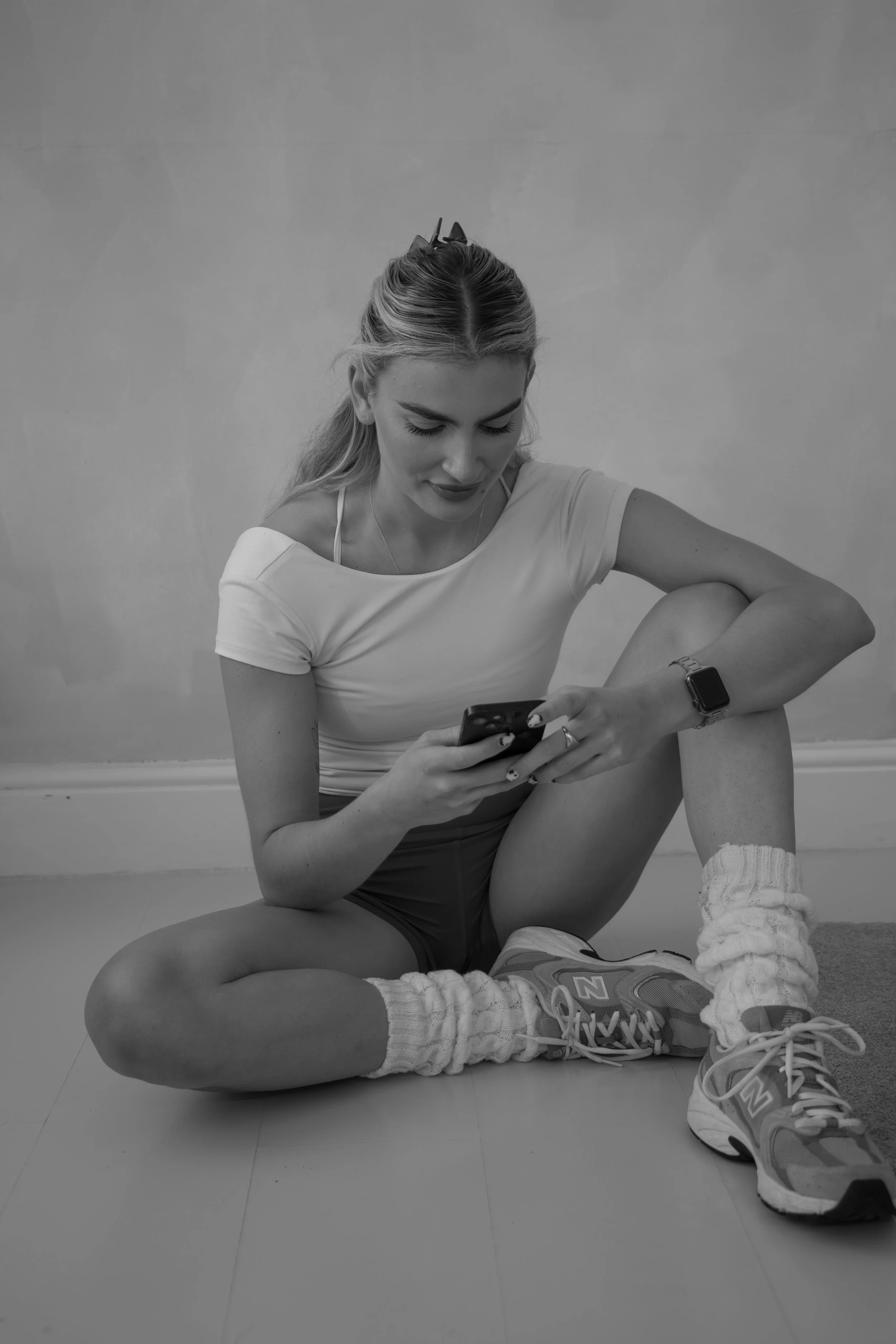 Young woman sitting on the floor with legs crossed, looking at her smartphone, wearing athletic clothing and sneakers, in black and white.