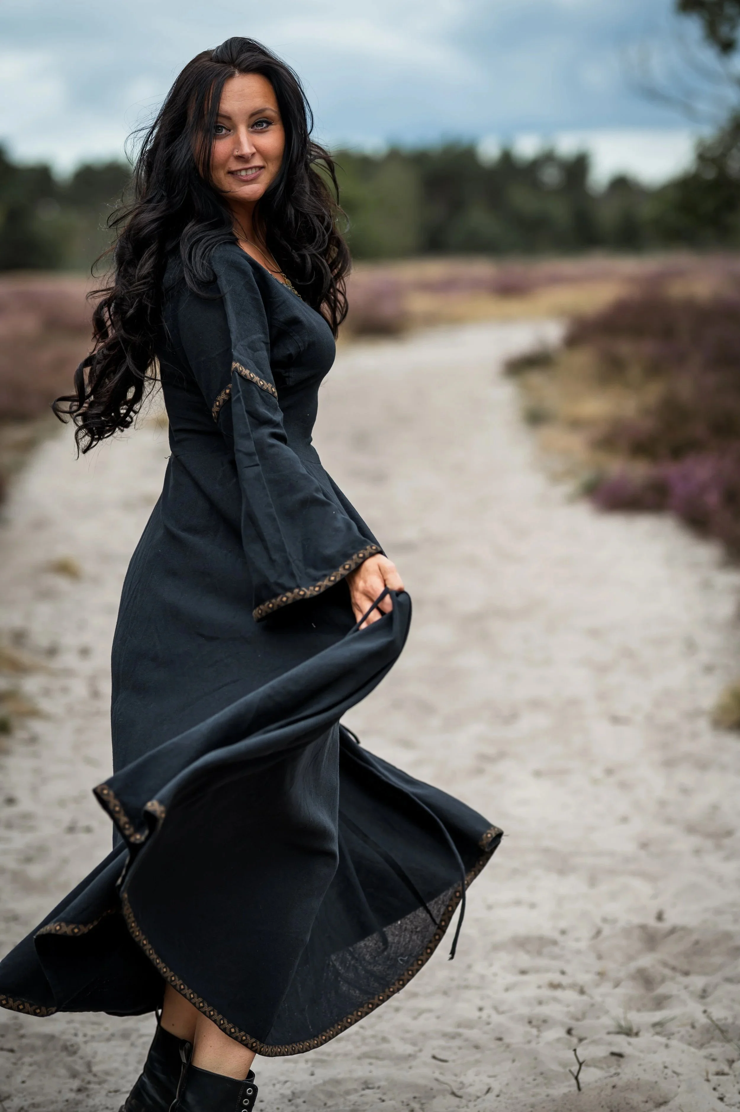 Een vrouw in een zwarte jurk staat op een zandpad in een natuurgebied, met een blauwe hemel en struiken of heide op de achtergrond.
