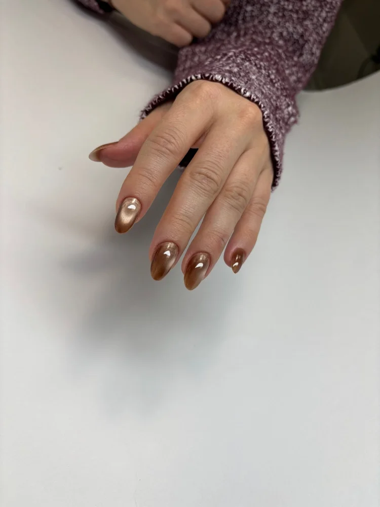 Close-up of a hand with metallic bronze-colored painted nails, resting on a white surface, with a person wearing a pink sweater in the background.
