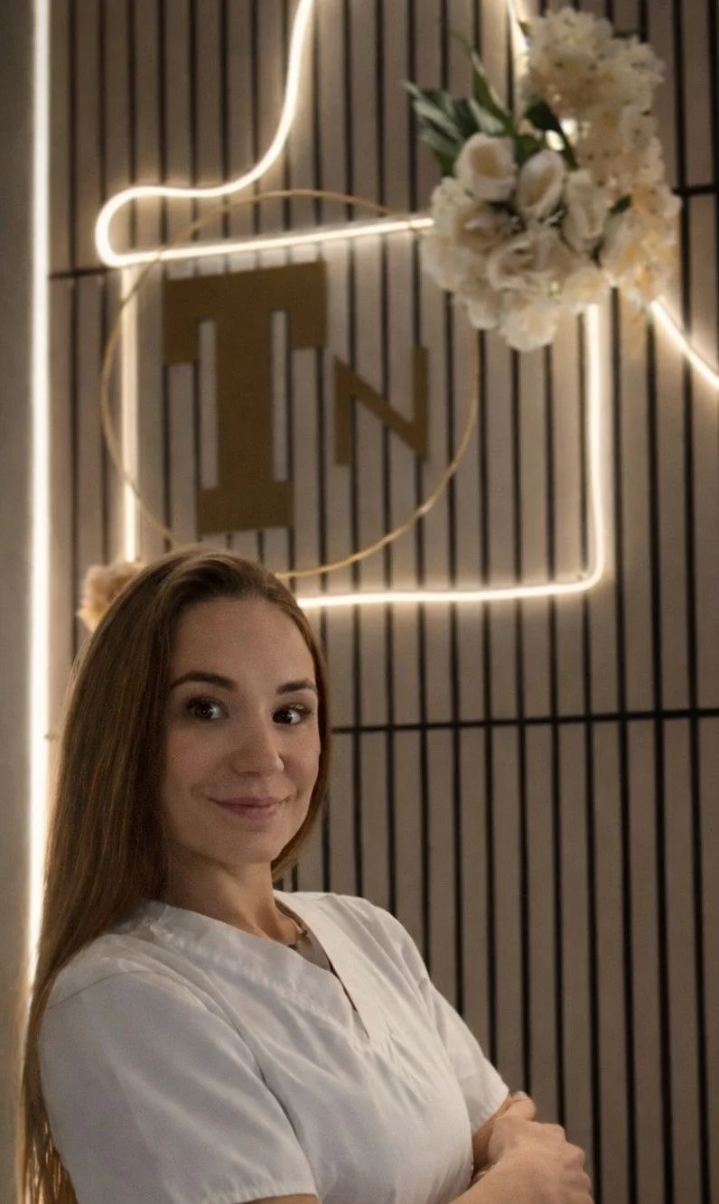 A woman with long brown hair smiling, wearing a white top, standing in front of a decorative wall with a neon light and a flower arrangement.