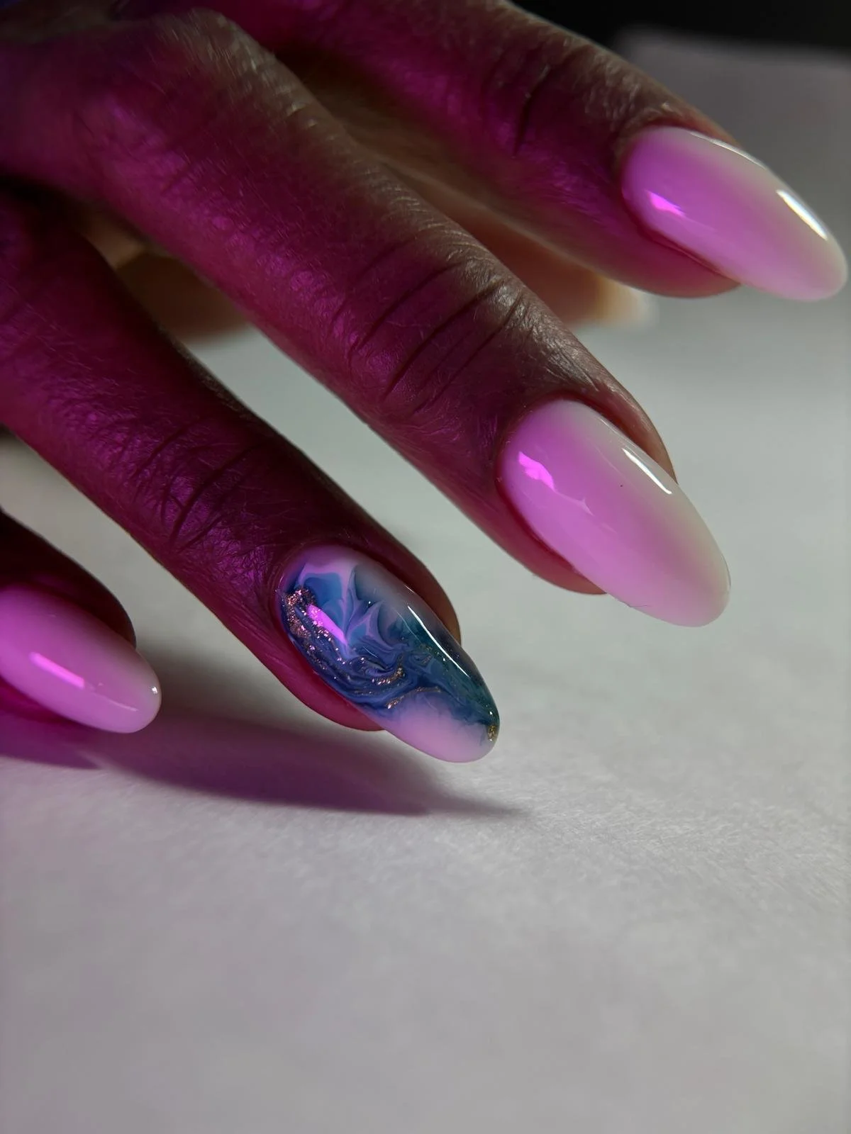 Close-up of a hand with pink and white metallic gradient nail polish, featuring one accent nail with blue, purple, and white marble design.