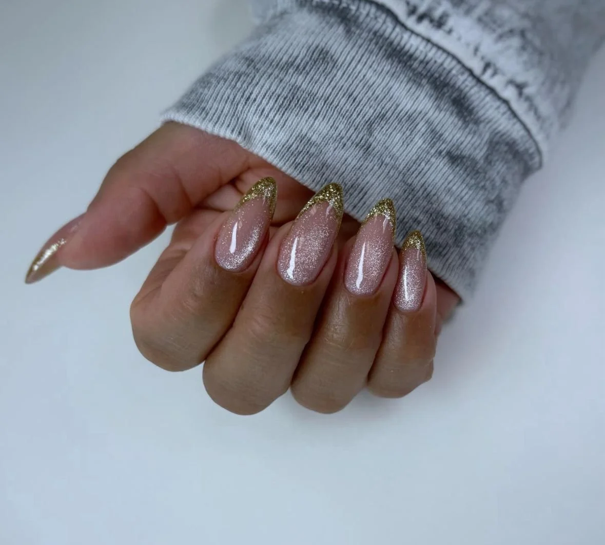 Close-up of a hand with decorated nails featuring a glittery, pinkish-nude polish with gold tips, close to a gray sweatshirt sleeve.