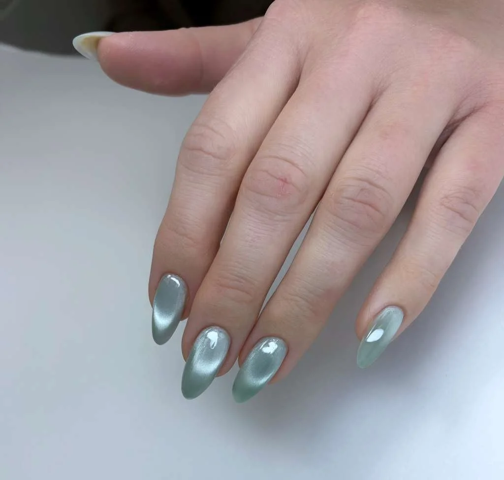 Hand with nails painted in a metallic silver-green color, resting on a white surface.