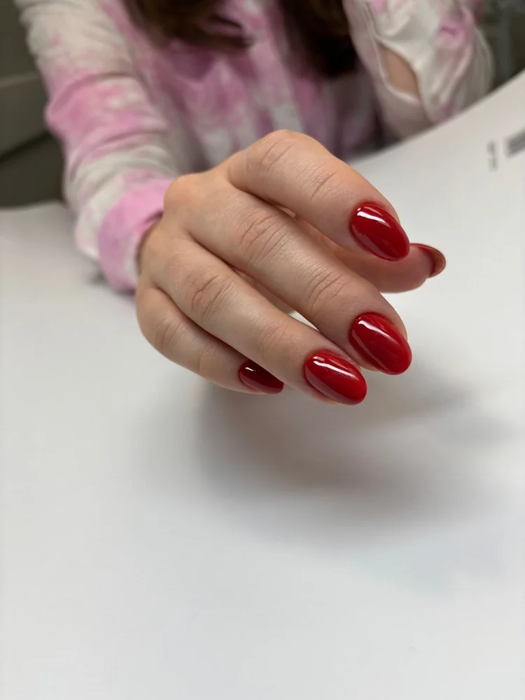 Close-up of a woman's hand with manicured red nails resting on a white surface, with part of a pink and white tie-dye long-sleeve shirt visible in the background.
