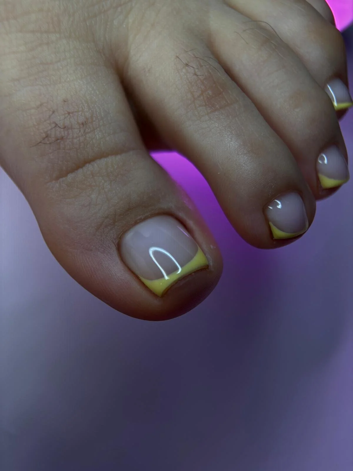 Close-up of fingers with freshly painted French manicure featuring yellow tips and clear gloss, against a purple background.
