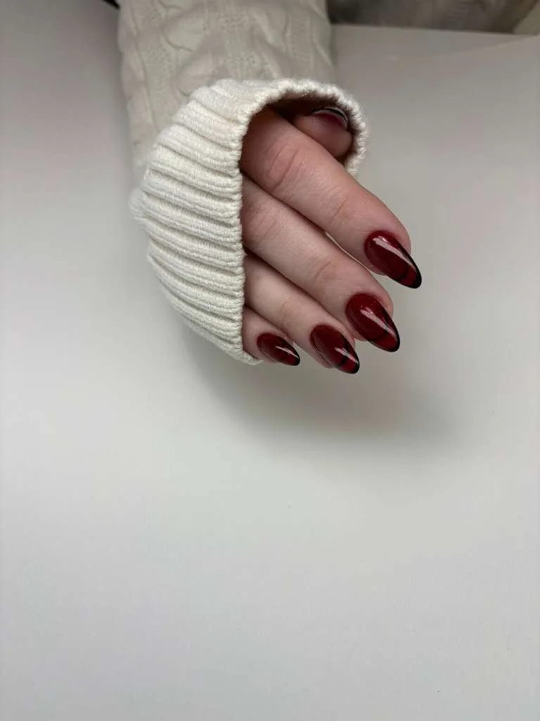 Close-up of a hand with long, almond-shaped nails painted in dark red, wearing a white knitted sweater sleeve.