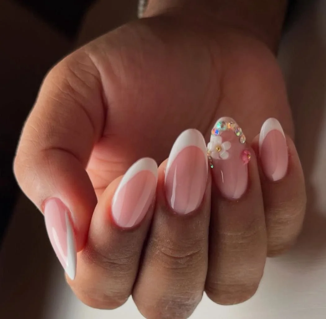 Close-up of a hand with manicured nails, featuring a French tip design with white tips and pinkish nail beds. The ring finger has decorative nail art with small rhinestones and a tiny white flower.