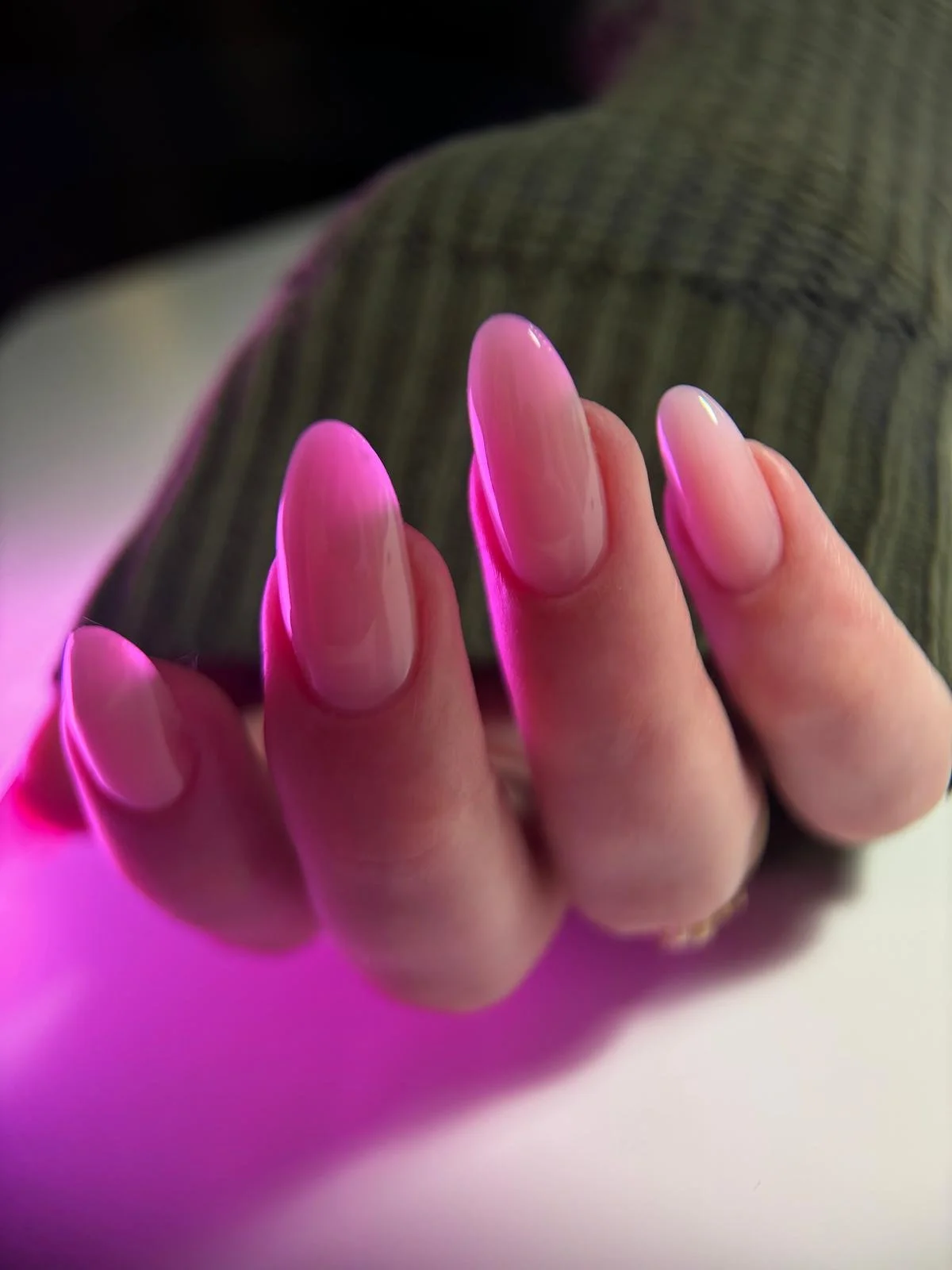 Close-up of a hand with long, oval-shaped, glossy pink nails, against a blurred background with pink and gray tones.