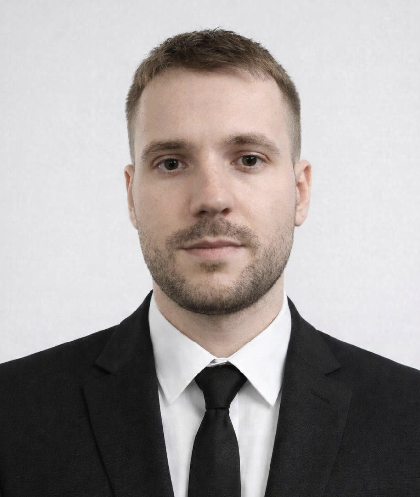 Siyah takım elbise ve kravat giymiş, düzgün saçlı ve sakallı genç adam portresi.