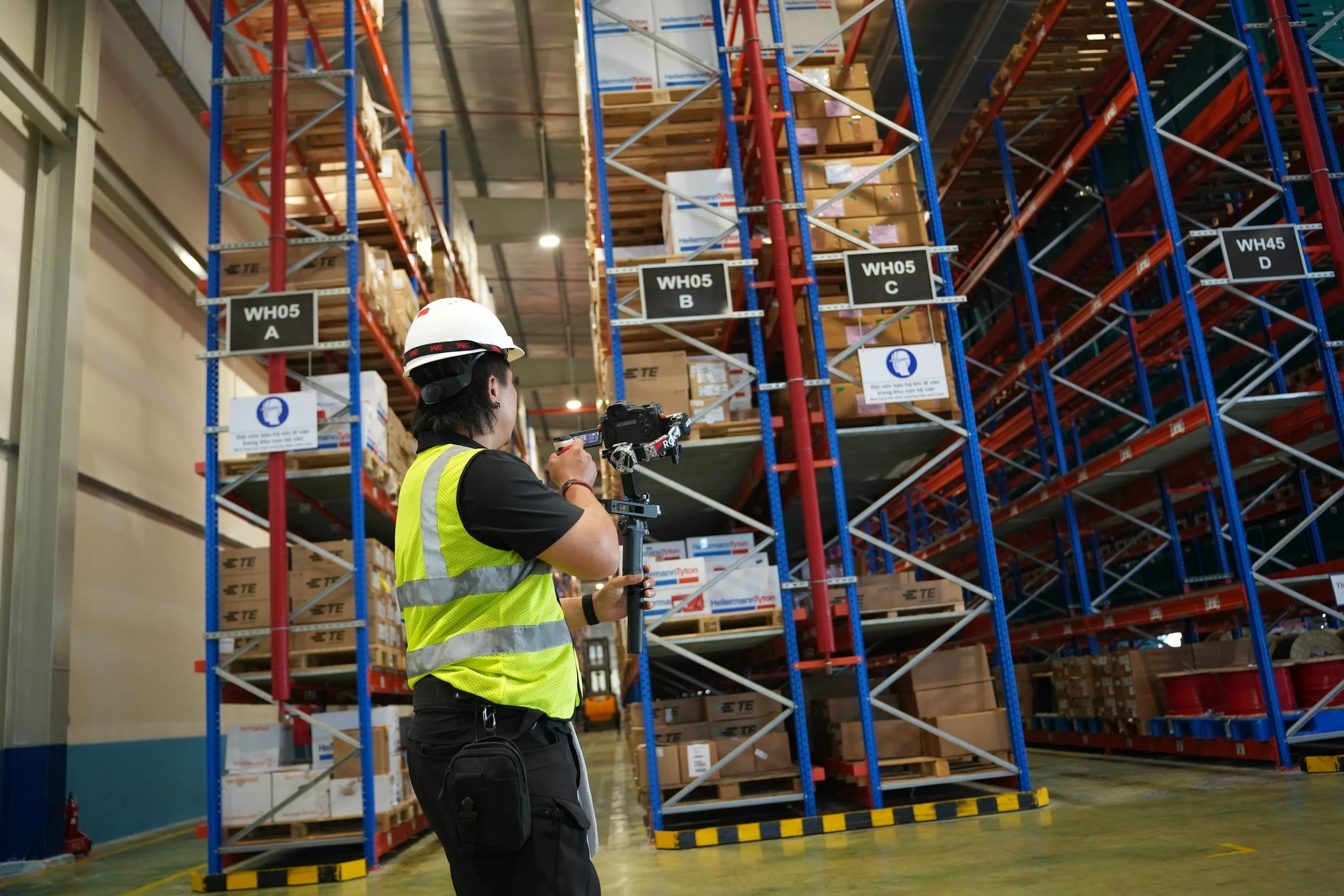 A worker wearing a safety helmet and a yellow safety vest holding a camera on a gimbal stabilizer inside a warehouse with tall shelves filled with boxes.
