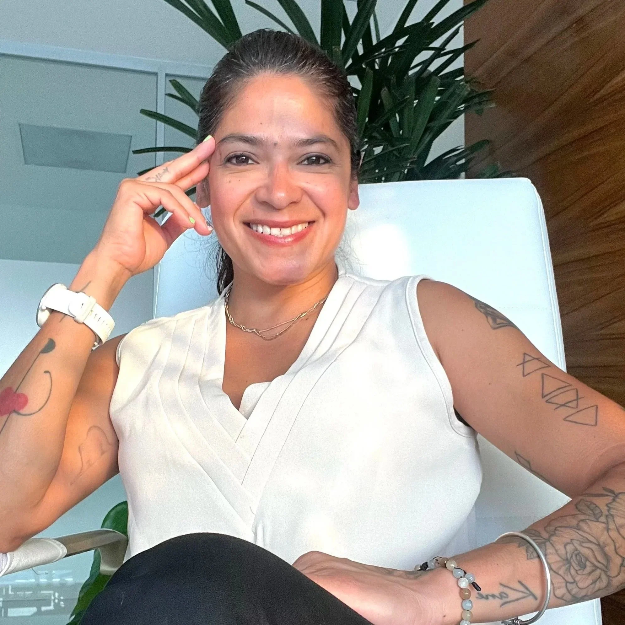 Mujer sonriendo, con cabello oscuro recogido en una coleta, llevando una camiseta blanca sin mangas y varias pulseras, sentada en una silla blanca en un ambiente moderno con plantas y paredes de madera.