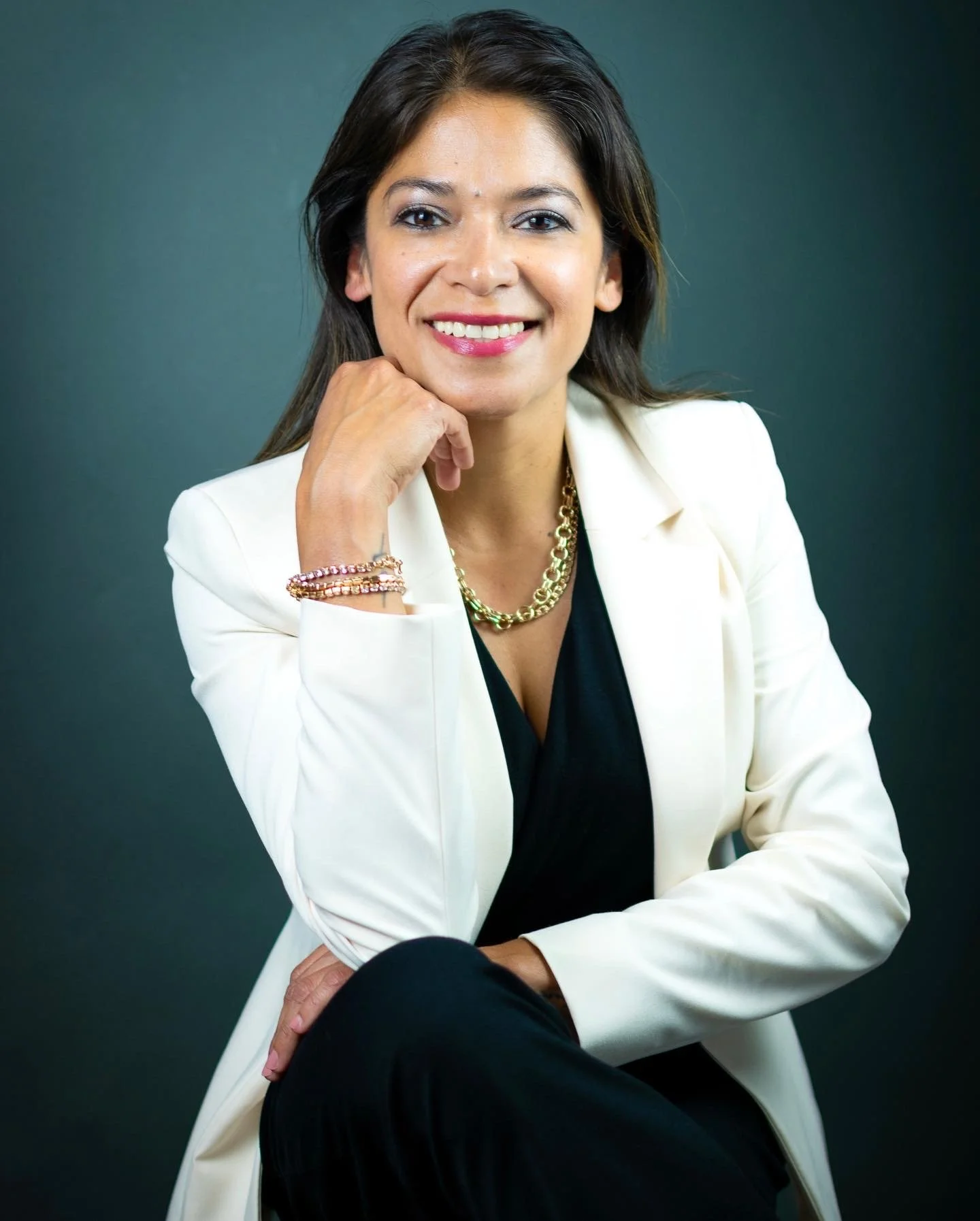 Mujer joven con cabello oscuro, piel clara y sonrisa, usando blusa negra, blazer blanco, collar dorado y pulseras, posando con un fondo gris oscuro.