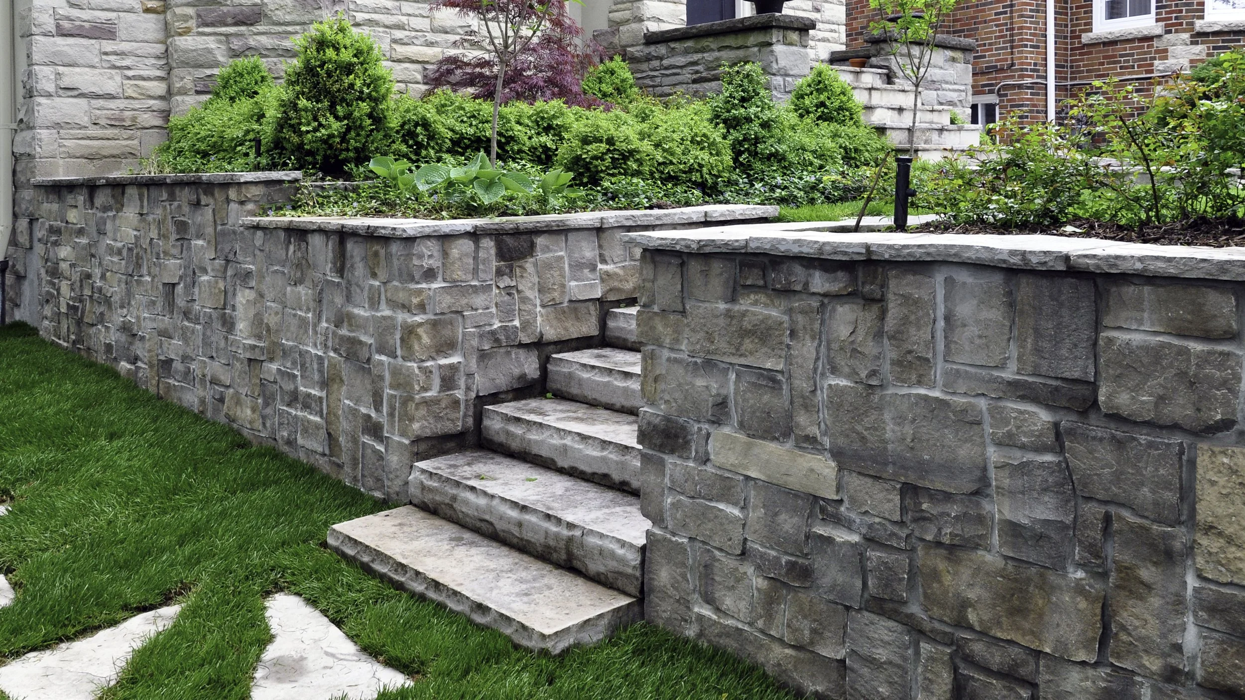 Stone retaining wall with steps leading up to a garden with green bushes and small plants, brick house in the background, and a well-manicured lawn with stone pathway in the foreground.