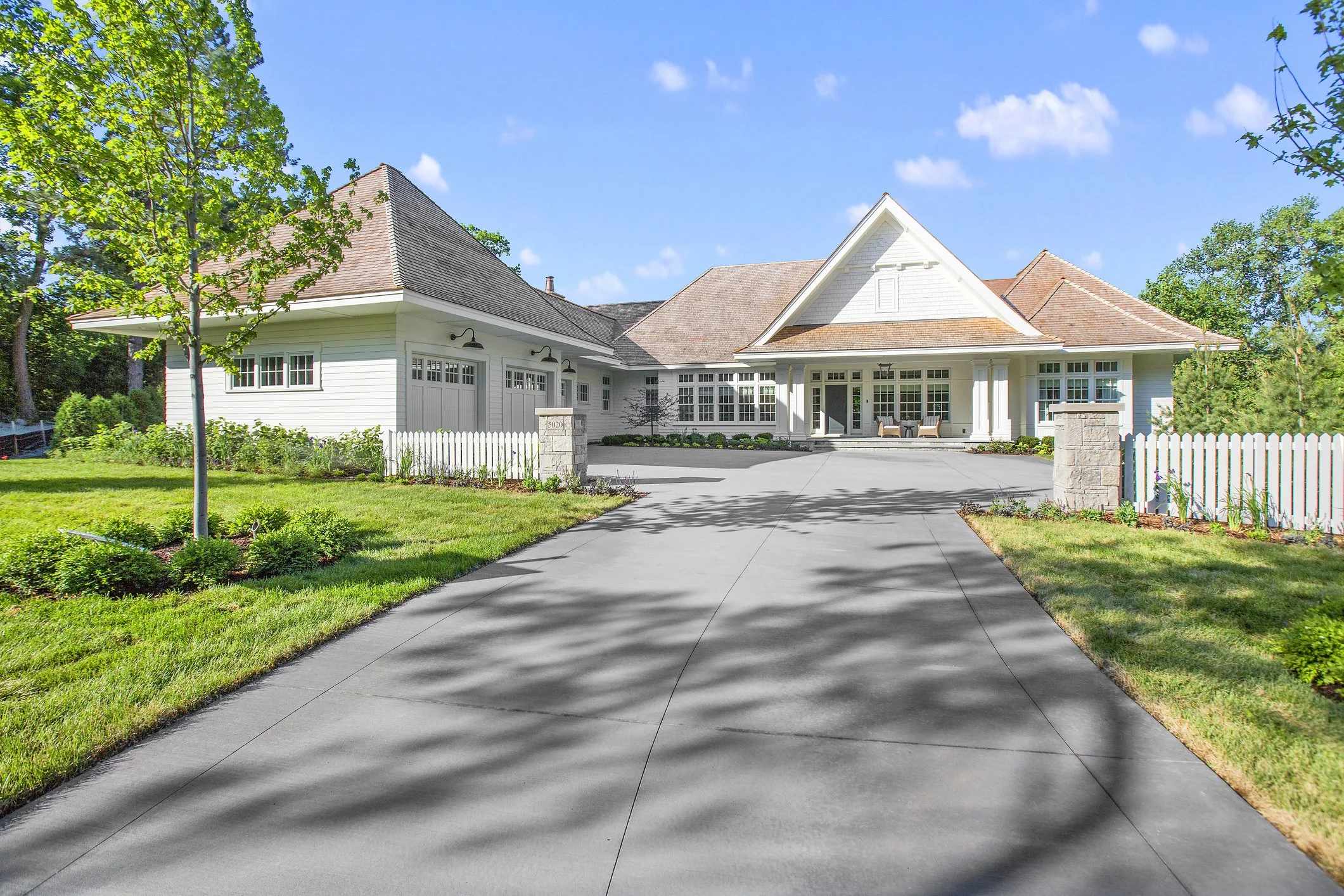A large white house with a driveway leading up to it, surrounded by green lawn and trees under a blue sky with clouds.