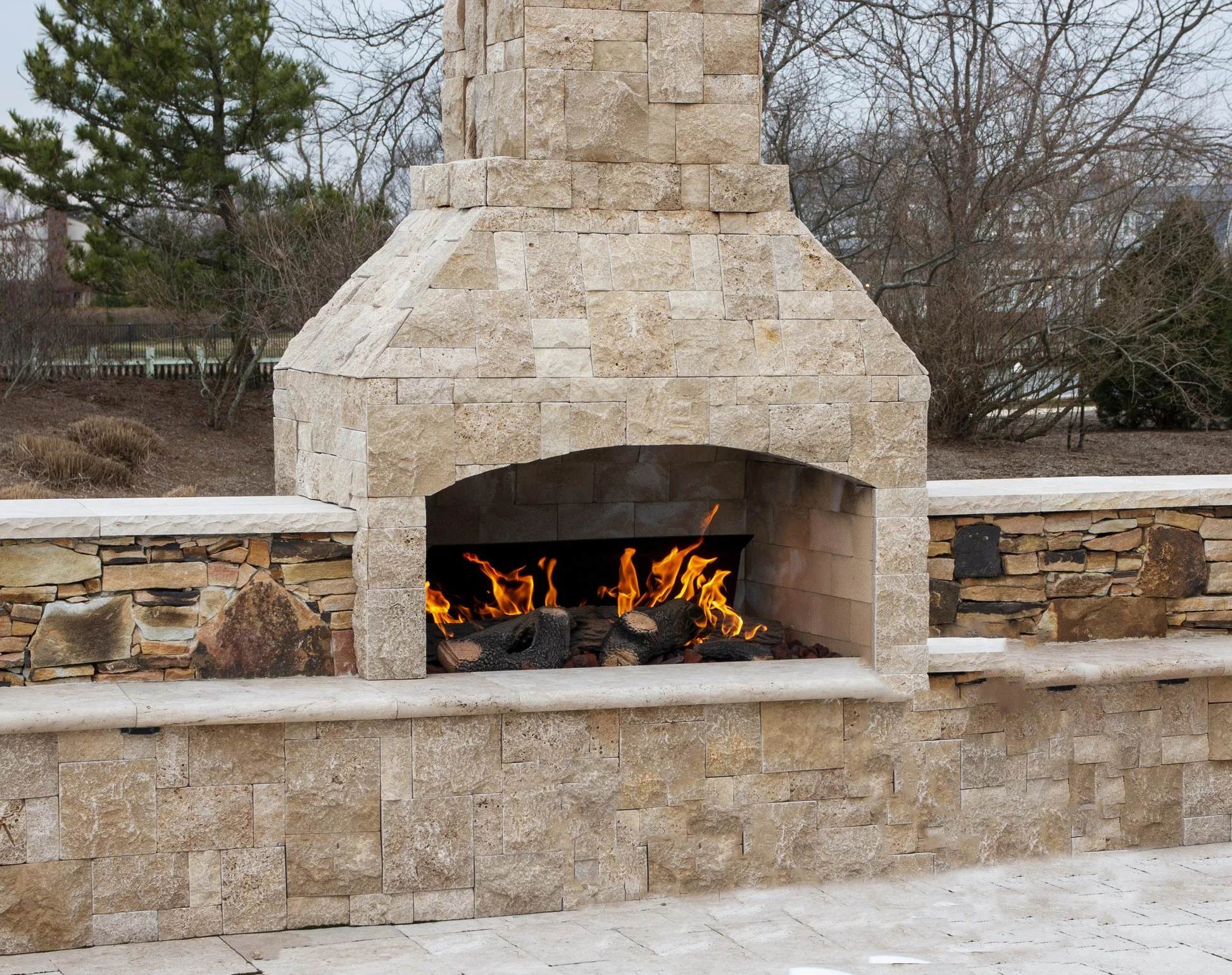 Outdoor stone fireplace with burning fire, surrounded by a stone and brick wall, with trees and a cloudy sky in the background.