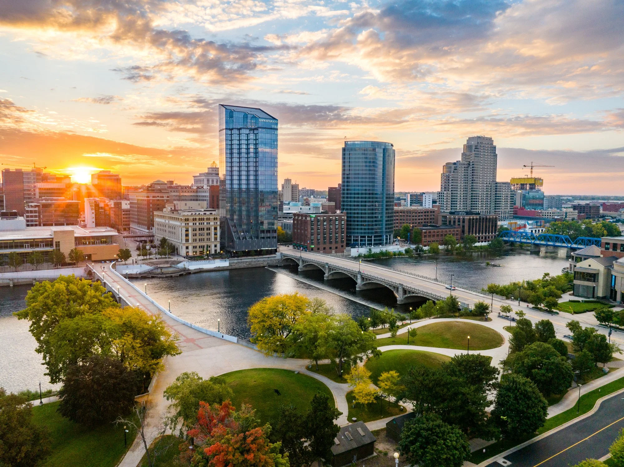 City skyline at sunset featuring modern high-rise buildings, a bridge over a river, and a park with walking paths and green trees in the foreground.