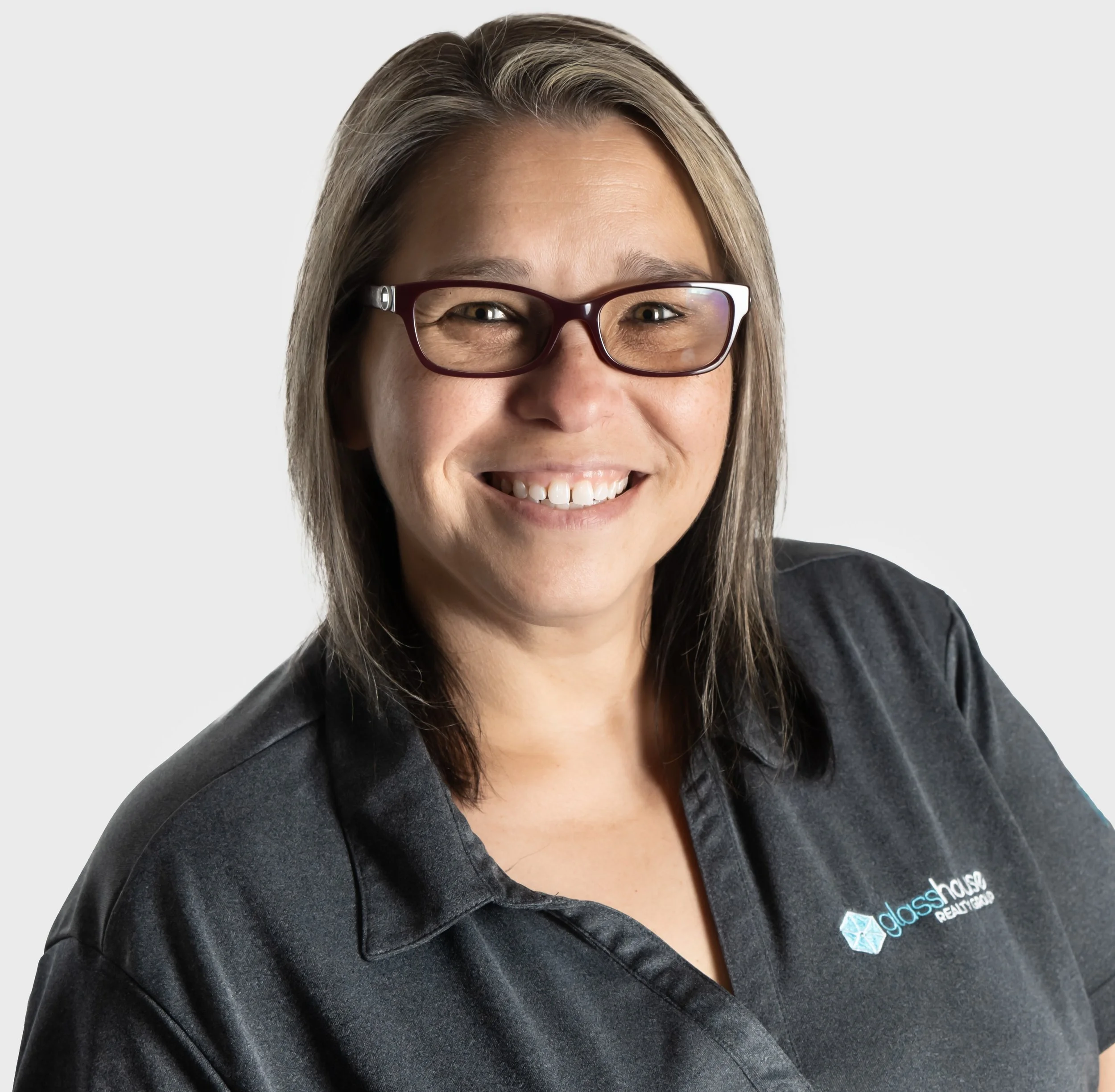 A woman with glasses and shoulder-length brown hair smiling against a plain white background, wearing a black polo shirt with a logo on the chest that says 'glass house'.