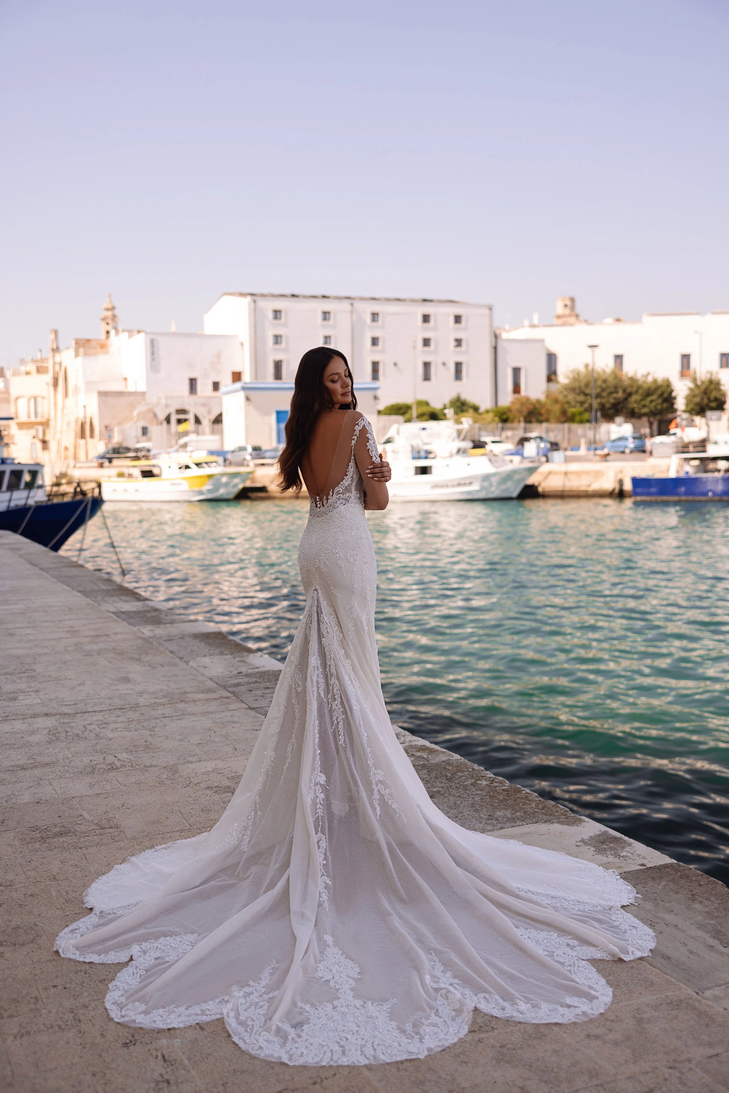 A bride in a white lace wedding dress with a train, standing on a stone dock by the water, with boats and a harbor in the background.