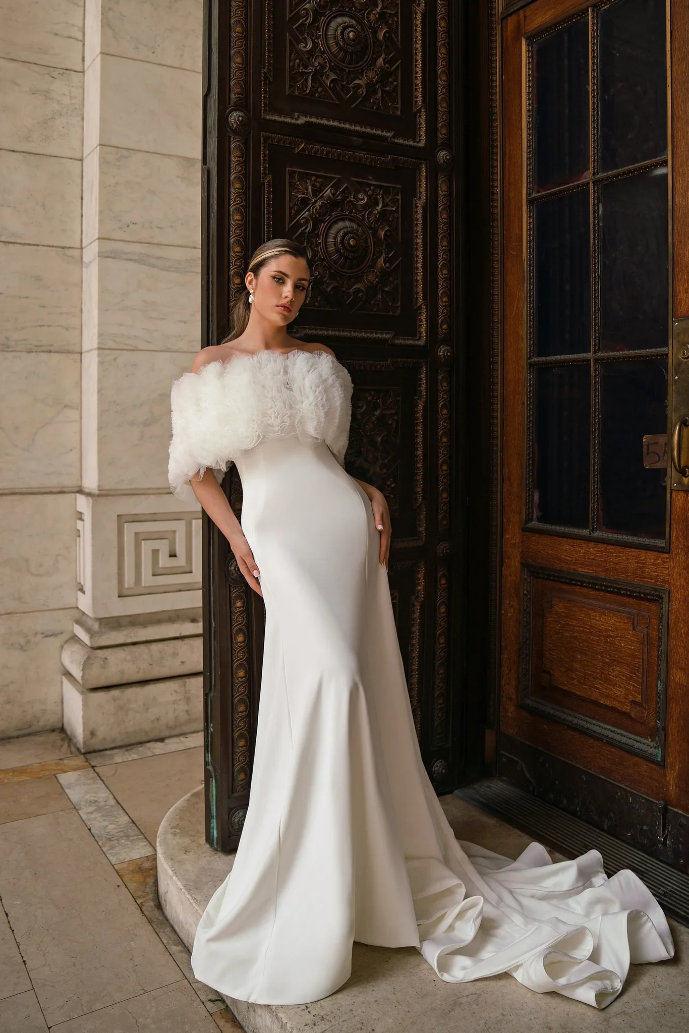 A woman wearing an elegant white off-the-shoulder gown with a feathered bodice standing in front of a large, ornate wooden door.