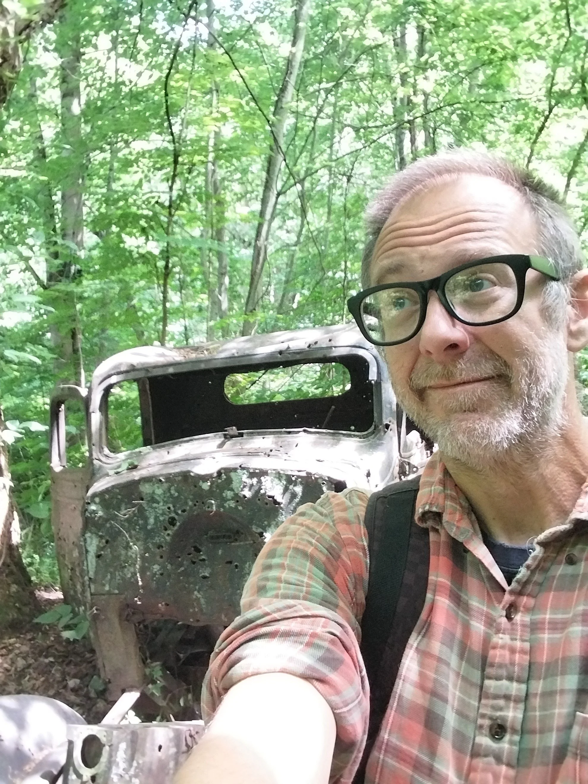 A man with glasses and a plaid shirt taking a selfie in a dense green forest, standing next to a rusty, old, burned-out vehicle.