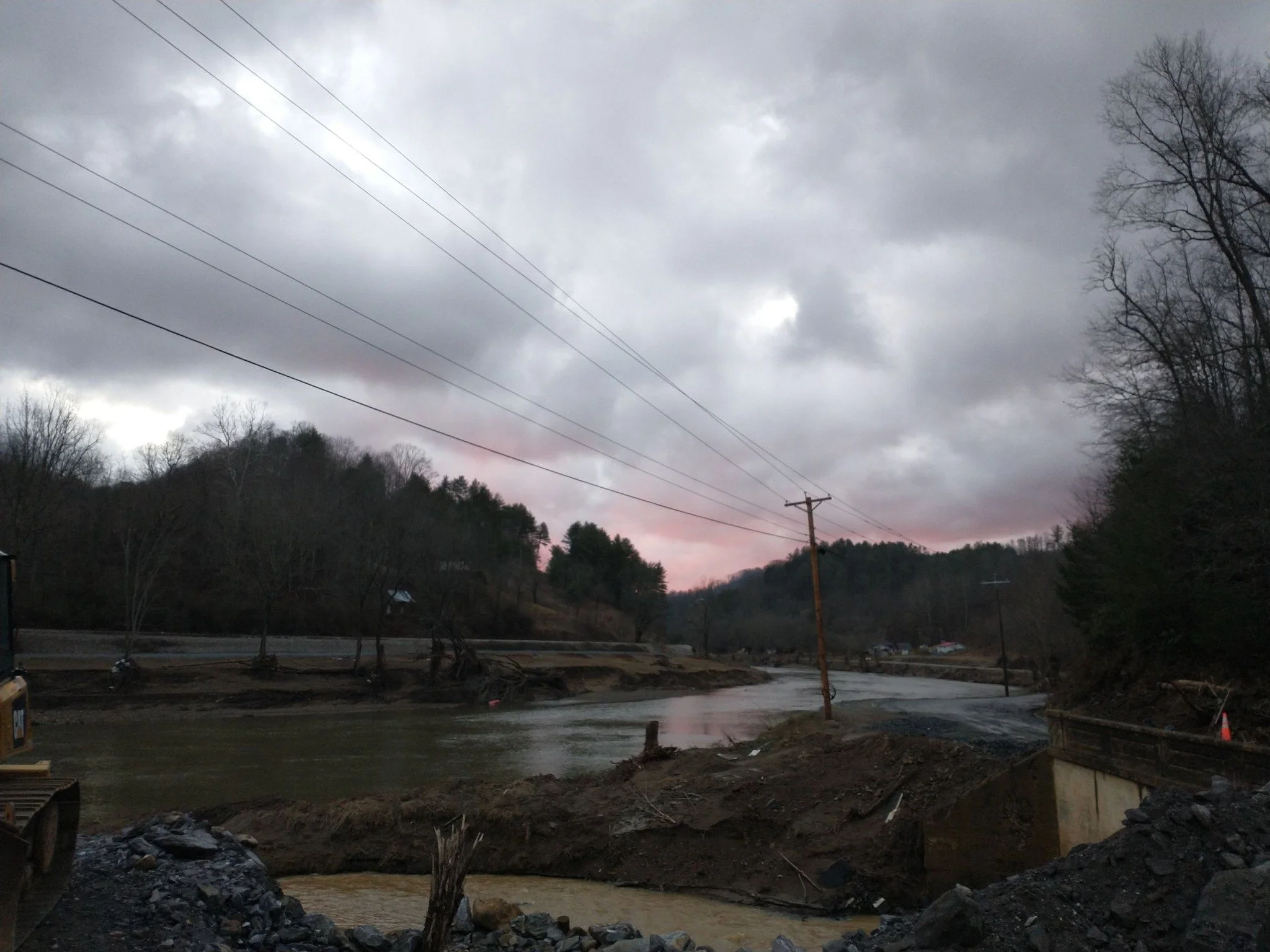 Overcast sky over a river with damaged banks, fallen trees, and damaged utility poles after a flood or storm, with construction equipment and debris in the foreground.