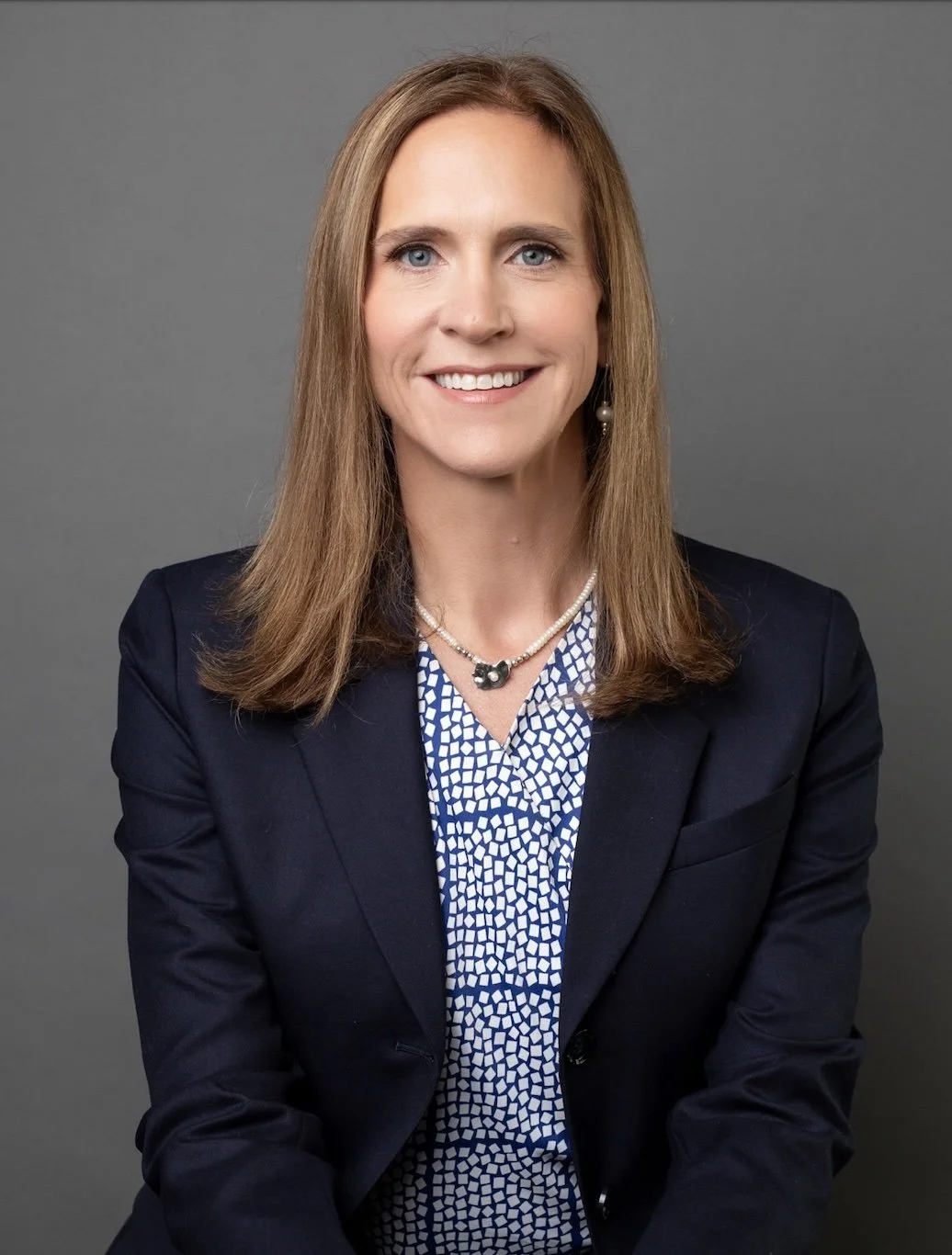 Professional woman with shoulder-length light brown hair smiling, wearing a dark blazer, patterned blouse, pearl necklace, and earrings, against a gray background.