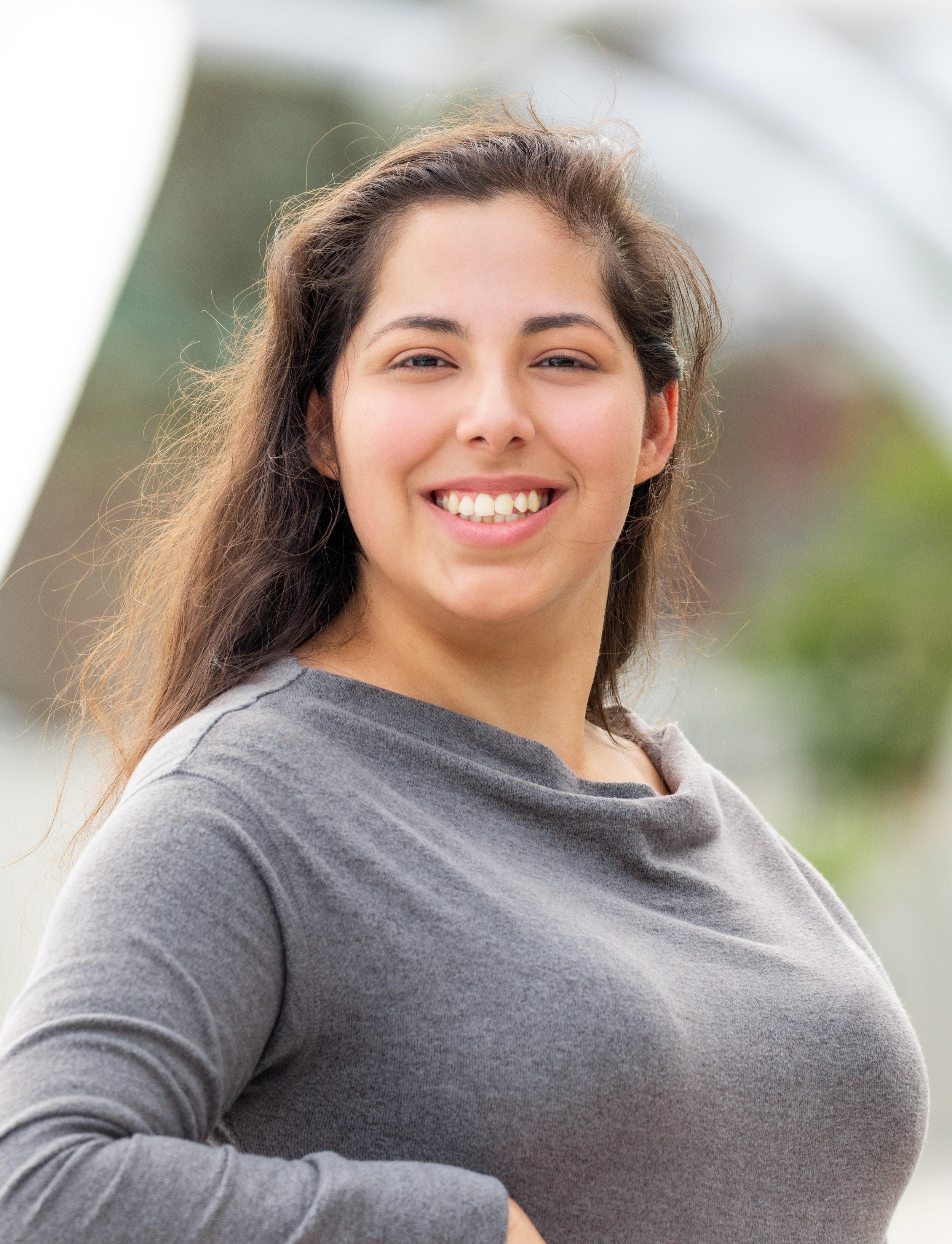 Smiling woman with long brown hair wearing a gray long sleeve shirt outdoors.