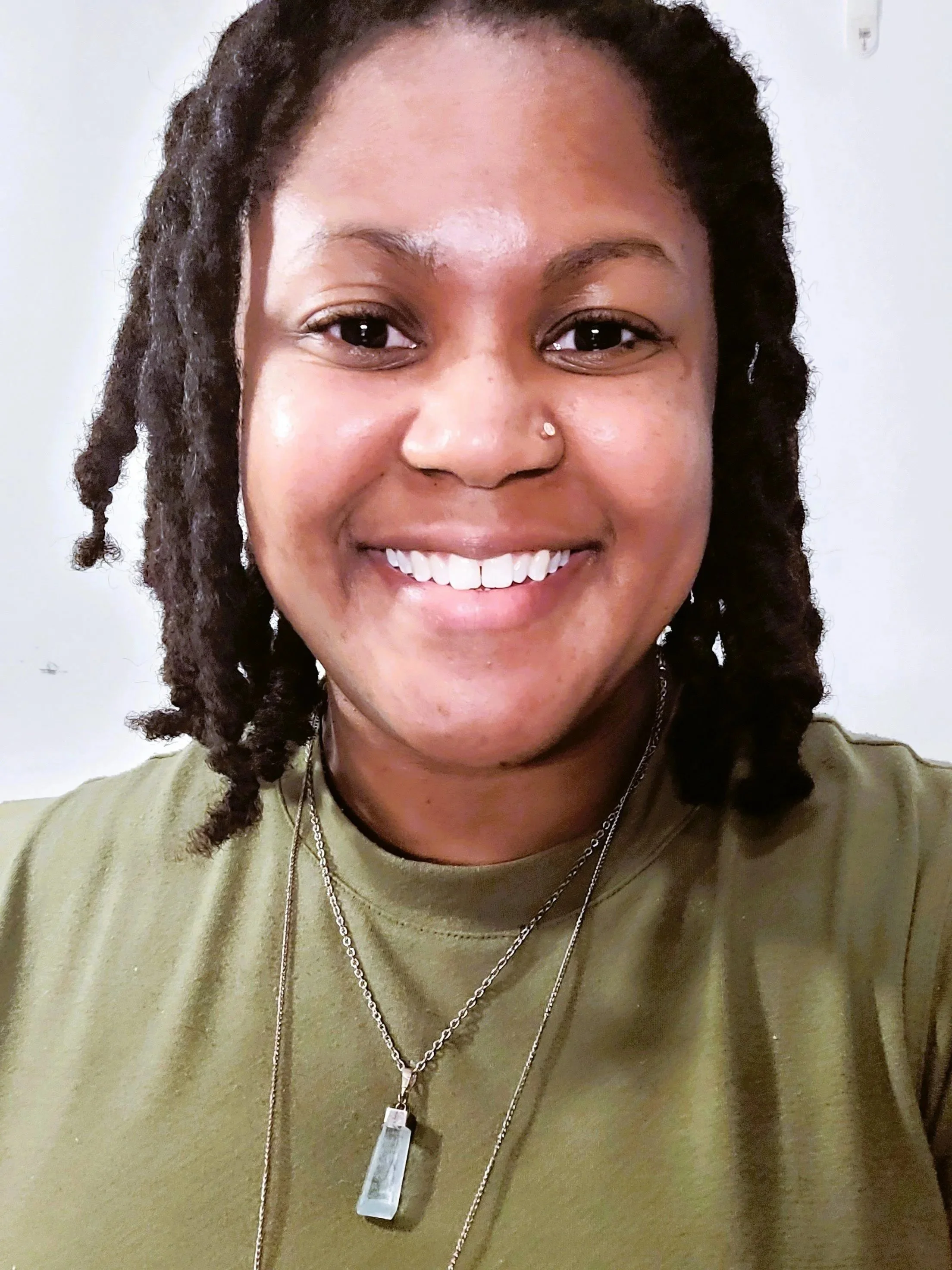 A smiling woman with dark brown, curly dreadlocks, wearing multiple necklaces and a nose piercing, dressed in an olive green shirt.