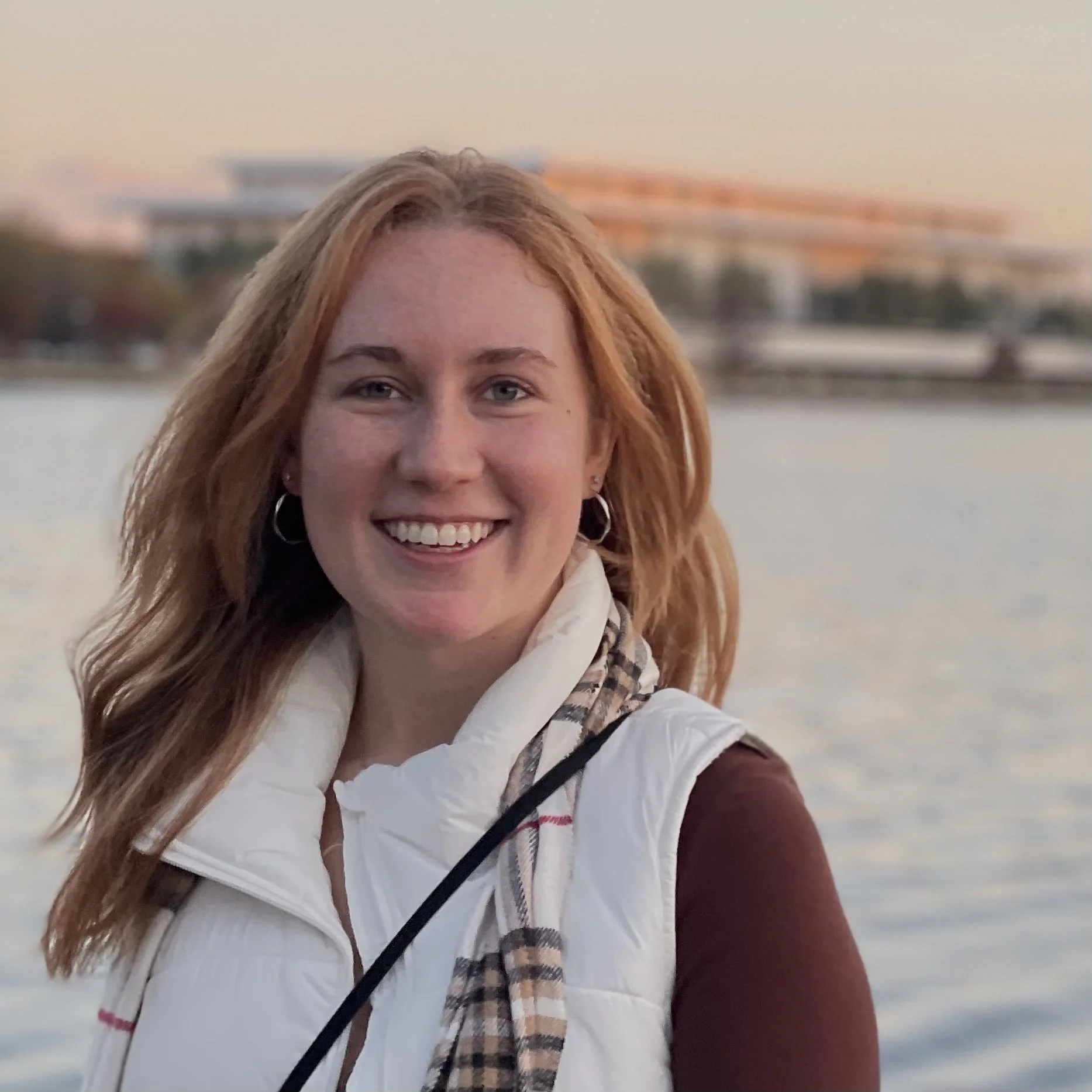 A woman with red hair smiling near a body of water at sunset, with a bridge and cityscape in the background.
