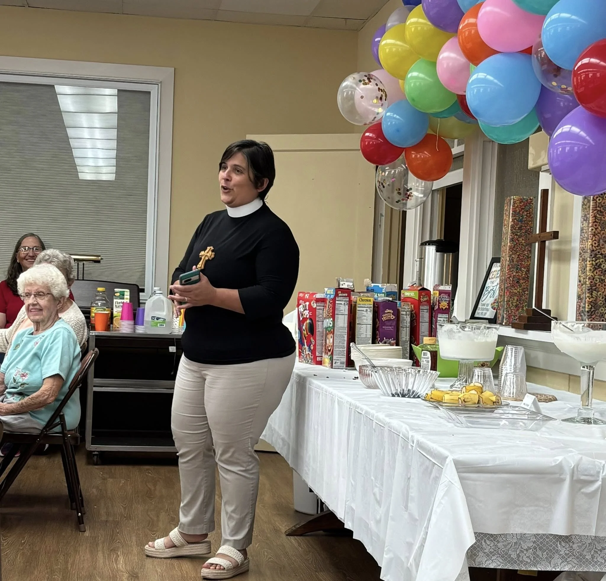 A woman pastor with a gold cross on her chest stands in front of a table with snacks and drinks, speaking to an audience in a room decorated with colorful balloons.